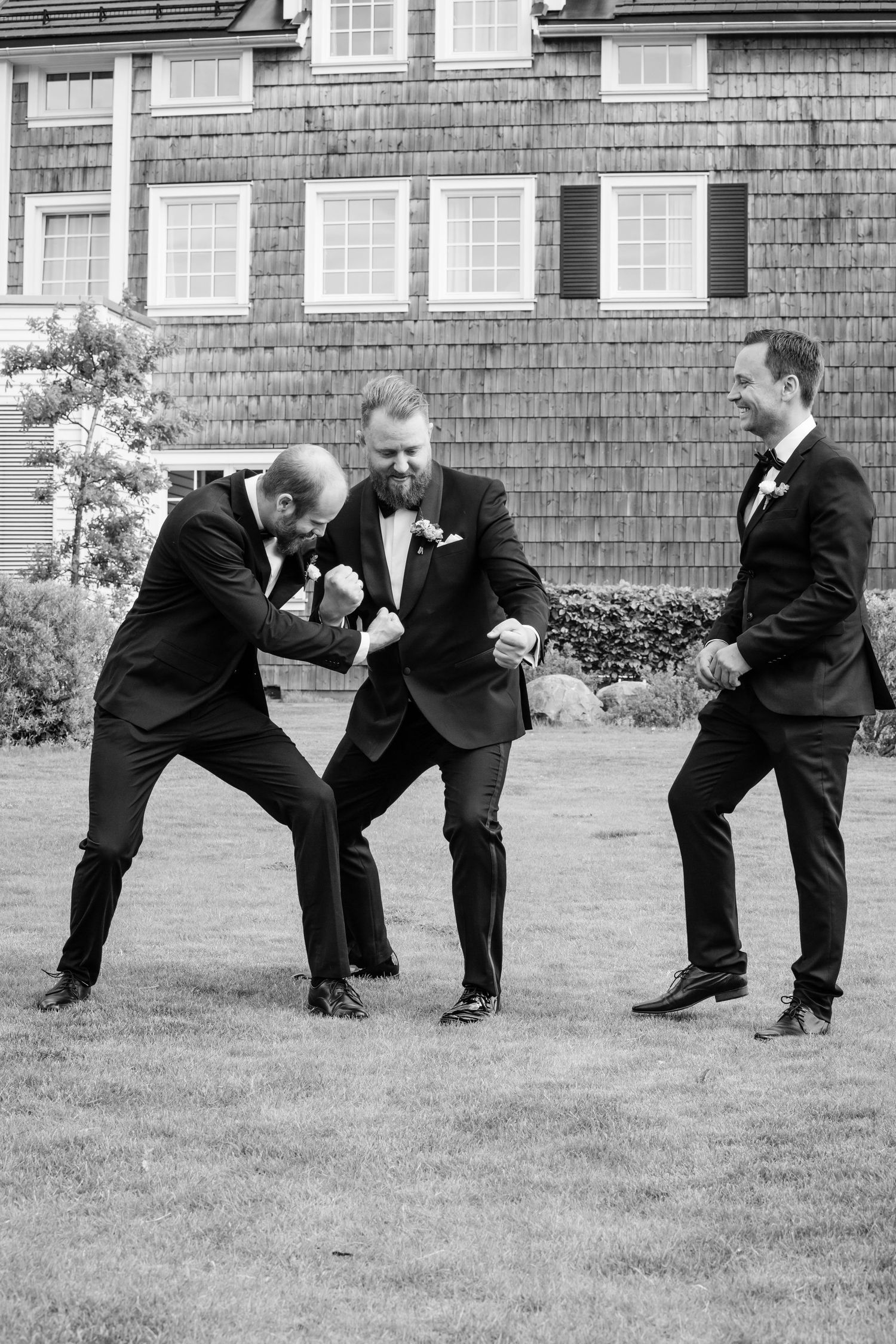 Groomsmen in black tuxedos performing silly poses on the lawn.