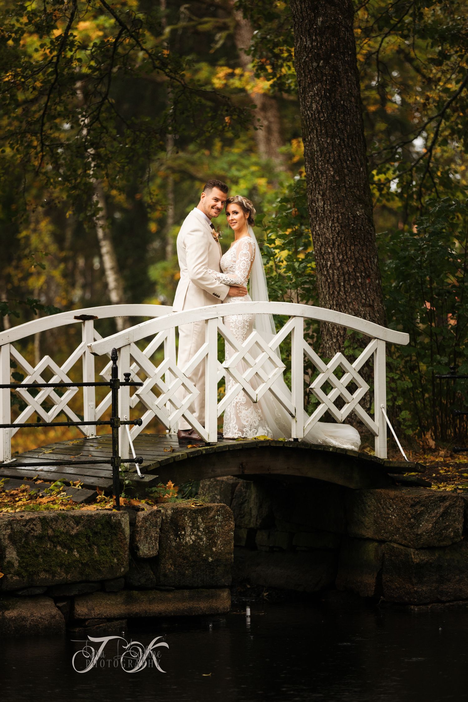 Newlyweds posing on white wooden bridge in scenic fall garden setting.