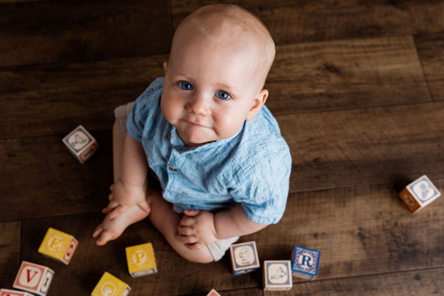 Baby in blue outfit playing with alphabet blocks on wooden floor.