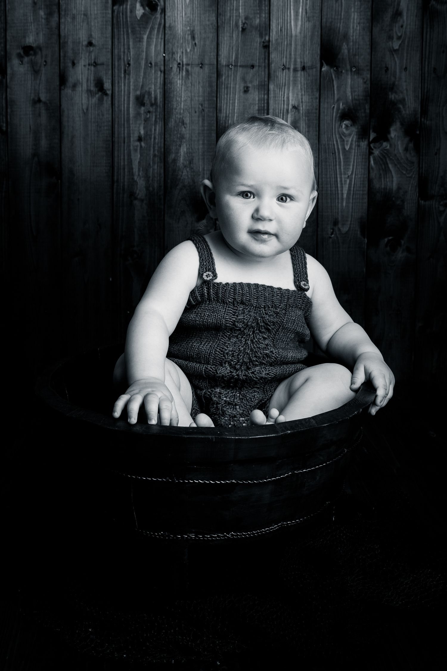 Black and white series showing baby in dark outfit sitting in vintage wooden bowl.