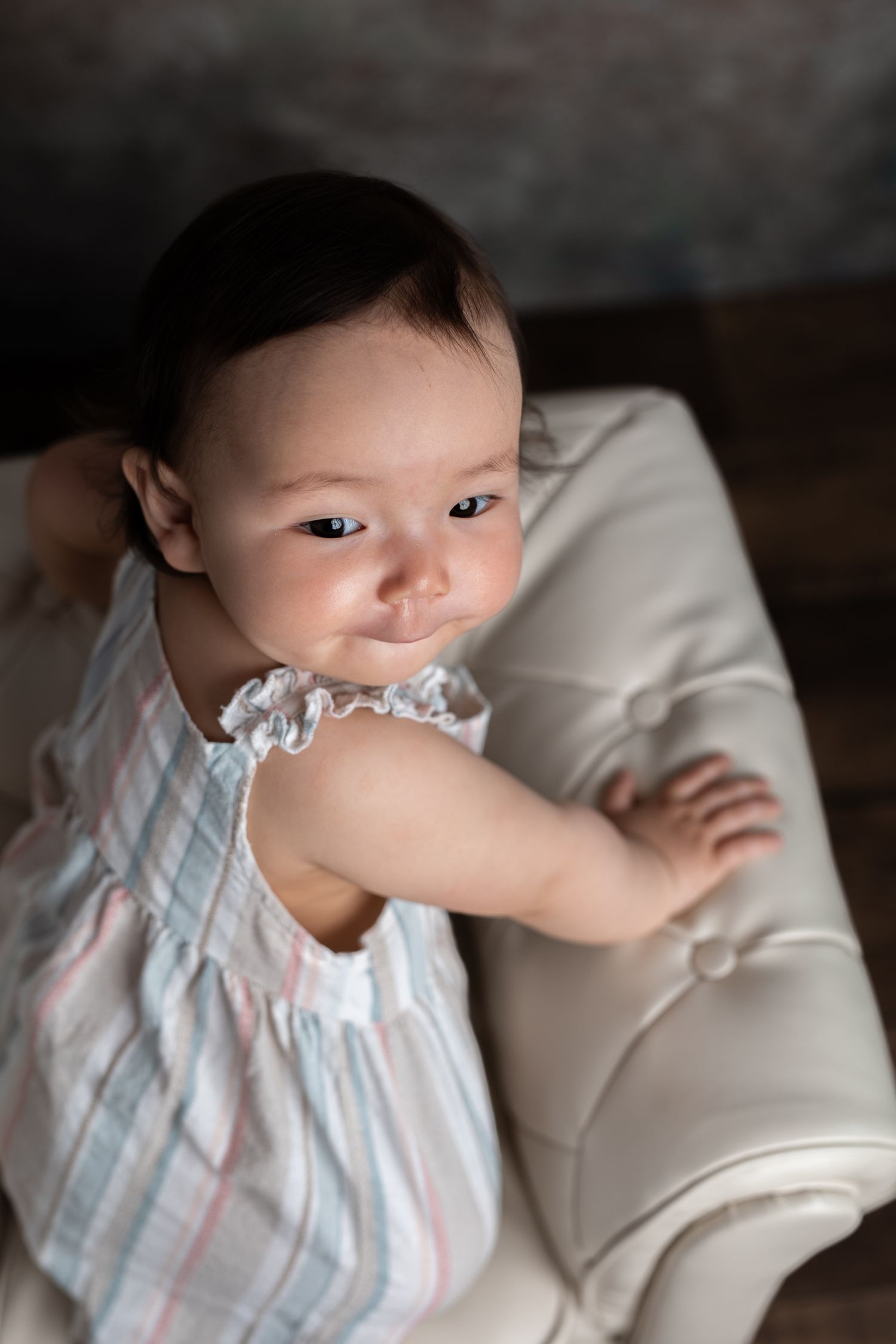 Baby in white dress sitting on cream couch during indoor photo shoot.