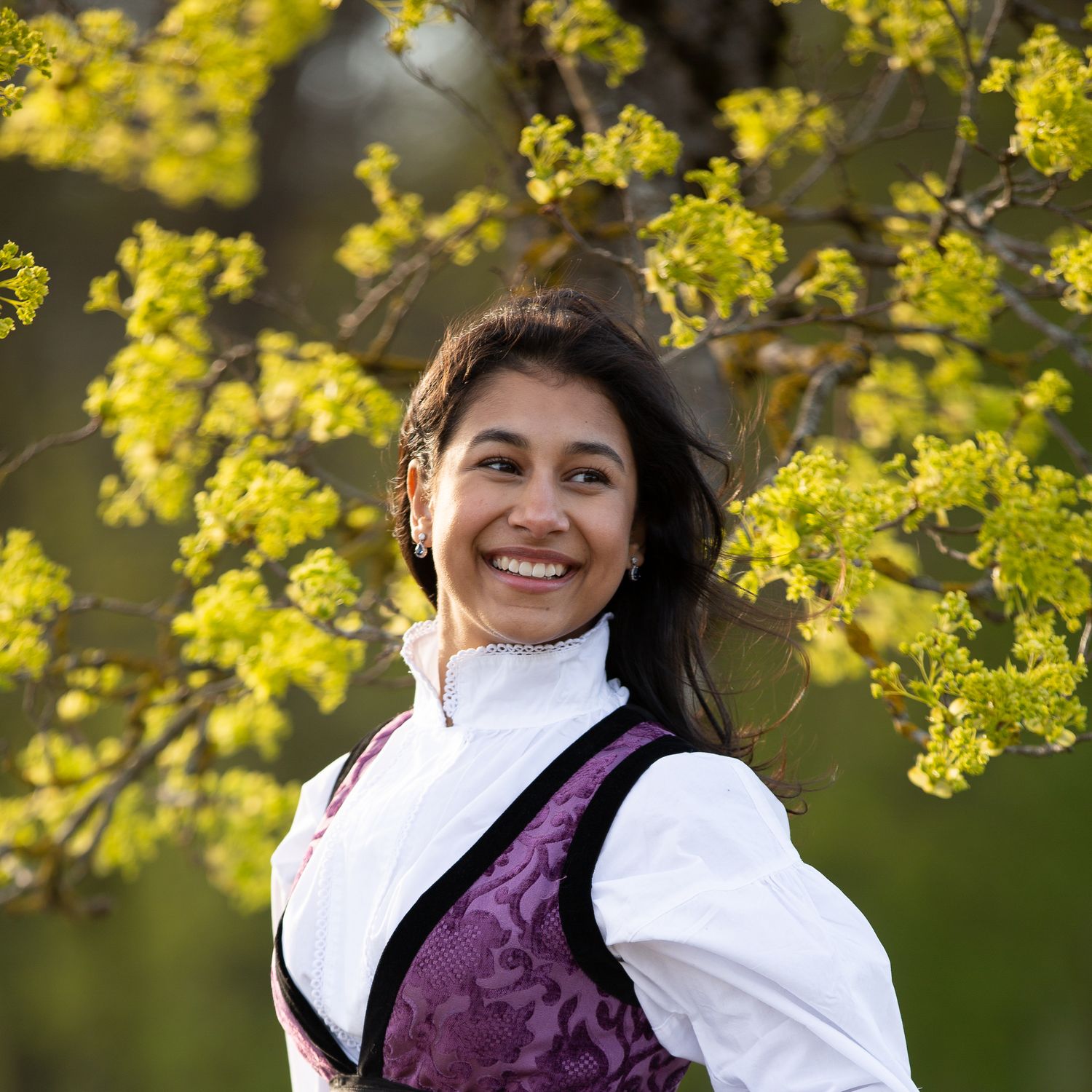 A smiling person wearing a white blouse and purple vest poses against yellow spring blossoms in nature.