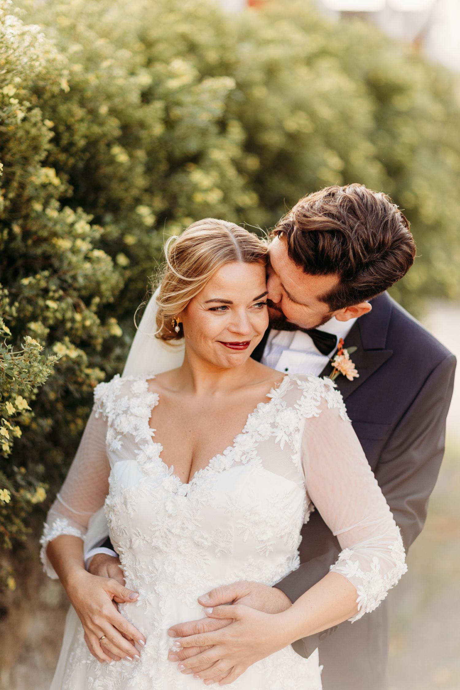 Wedding couple embracing outdoors with lace dress and natural greenery backdrop.