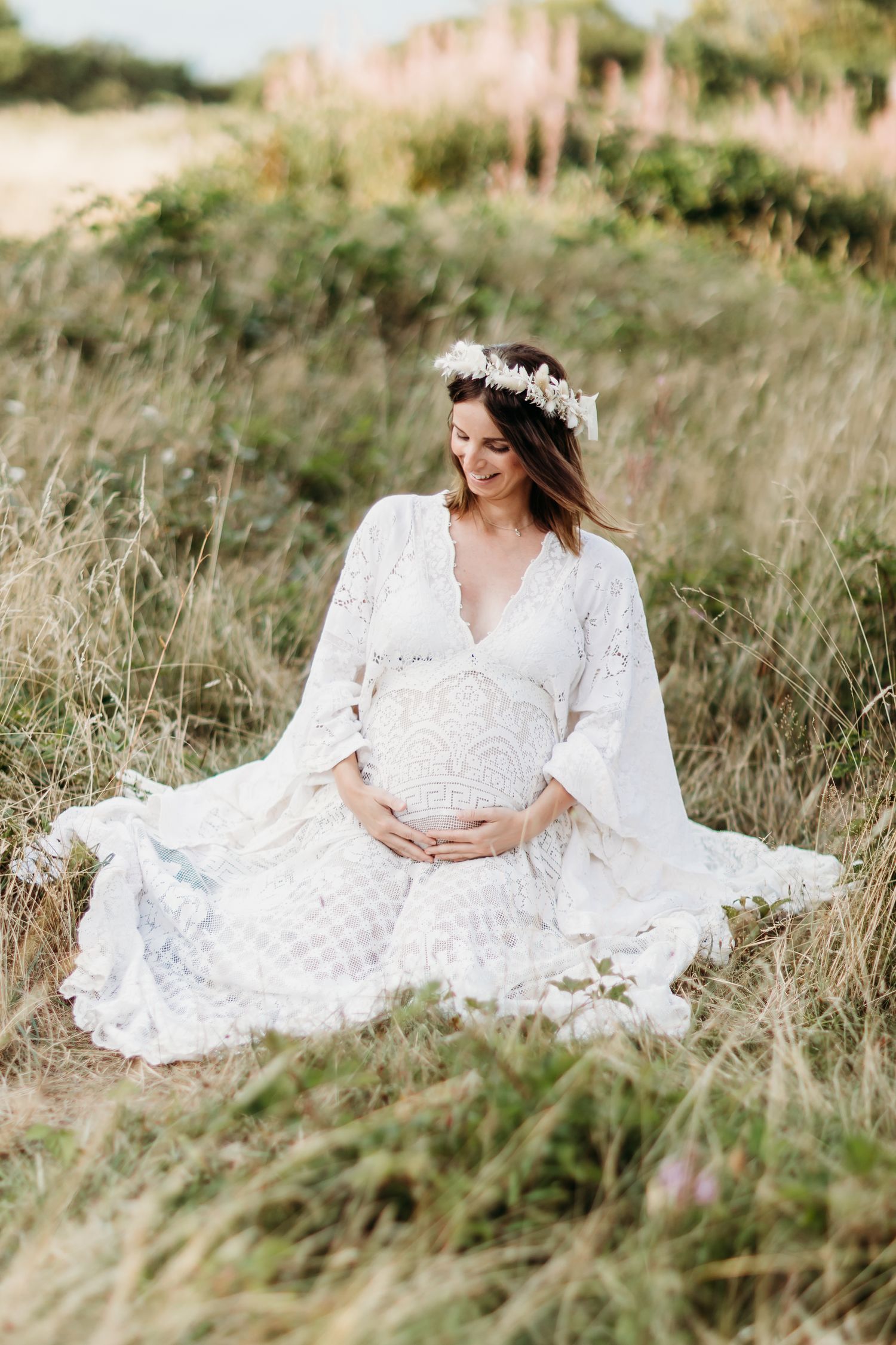 Woman in white lace dress poses gracefully in a sunlit meadow of tall grass.