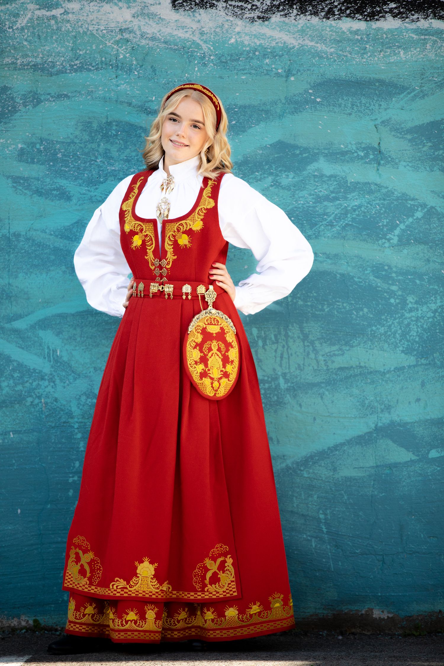 Red and gold Norwegian bunad dress photographed against turquoise wall.
