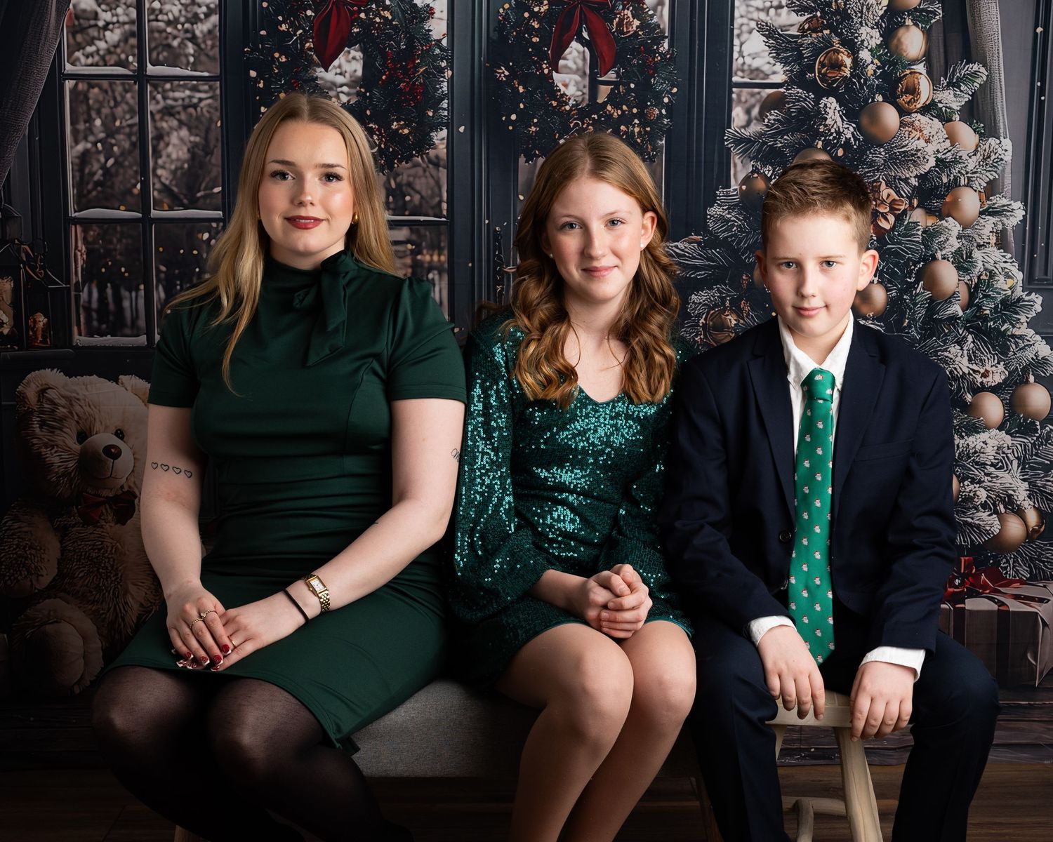 Three siblings in green holiday outfits pose together in front of a decorated Christmas tree for a festive family portrait.
