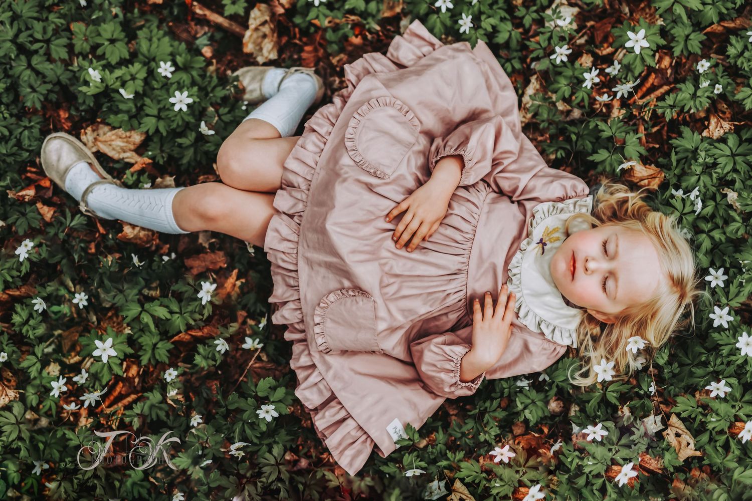 A child in a pink ruffled dress lies peacefully among white flowers and greenery.