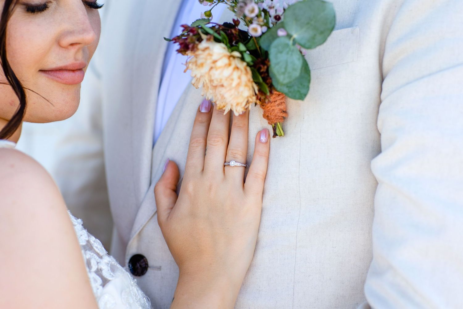 Close up detail of bride's hand with ring against groom's white suit jacket with peach boutonniere.