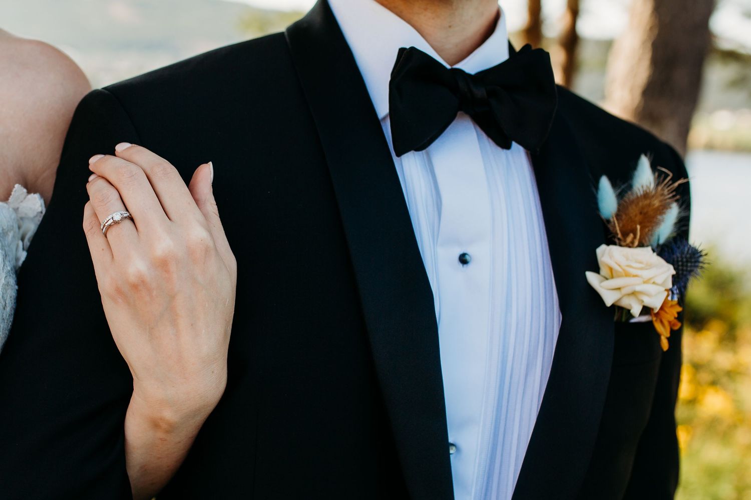 Close-up detail of a groom's tuxedo, boutonniere and bride's hand showing wedding ring.