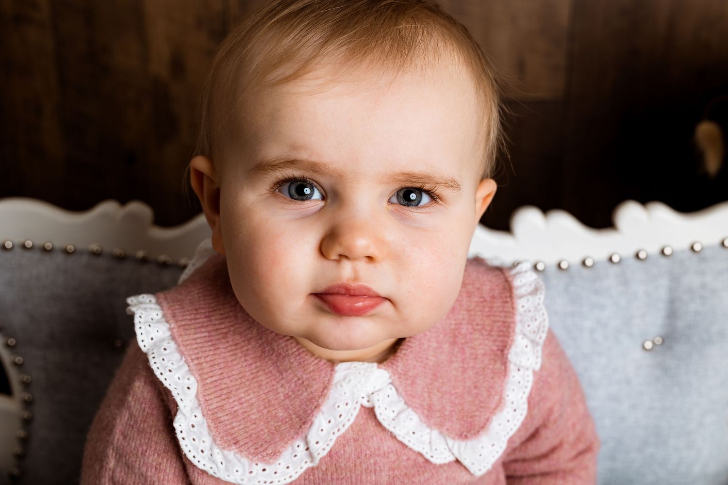 Baby wearing pink dress with white collar lying on wooden floor in natural light portrait.
