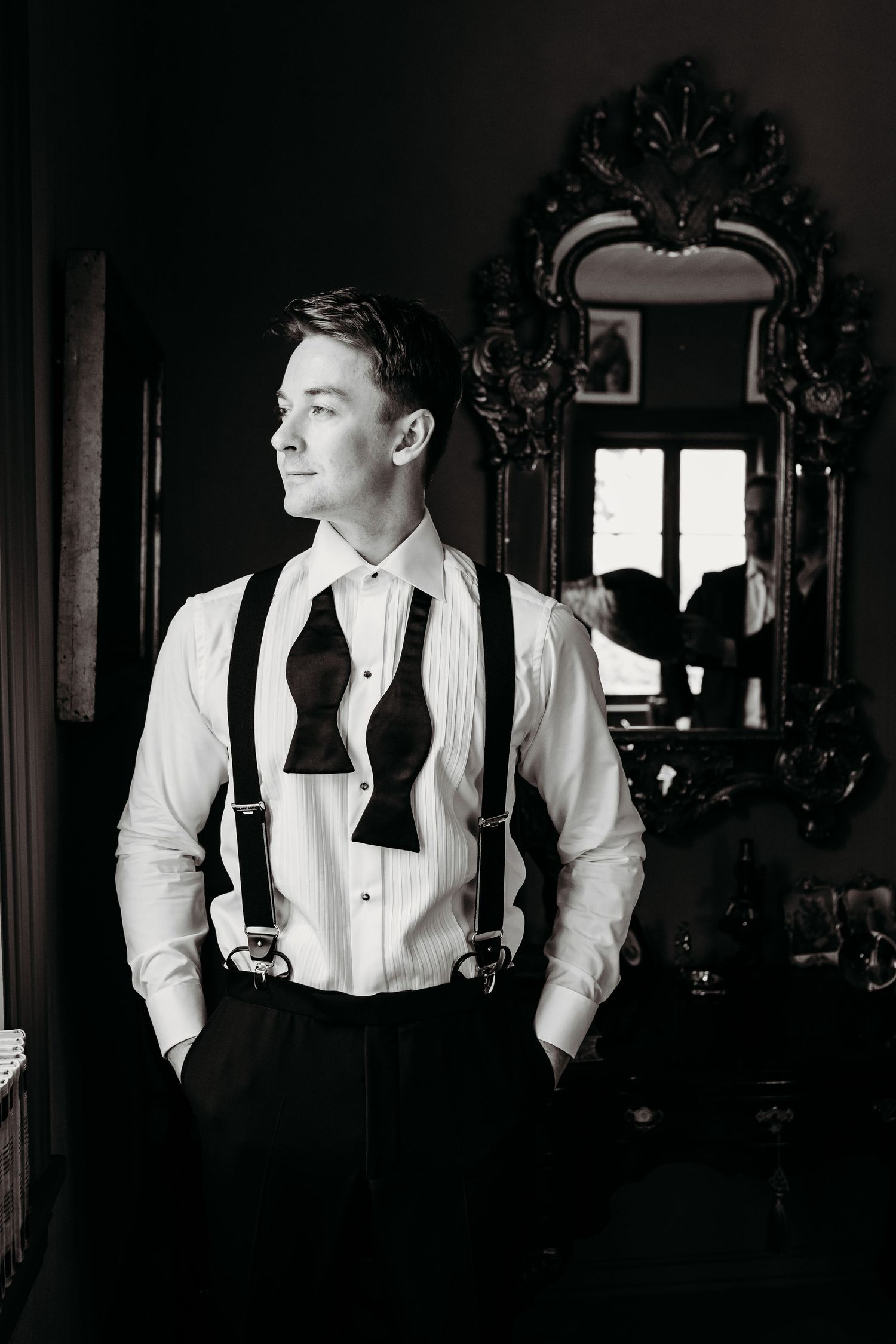 Classic black and white portrait of a groom in suspenders and dress shirt standing by an ornate mirror.