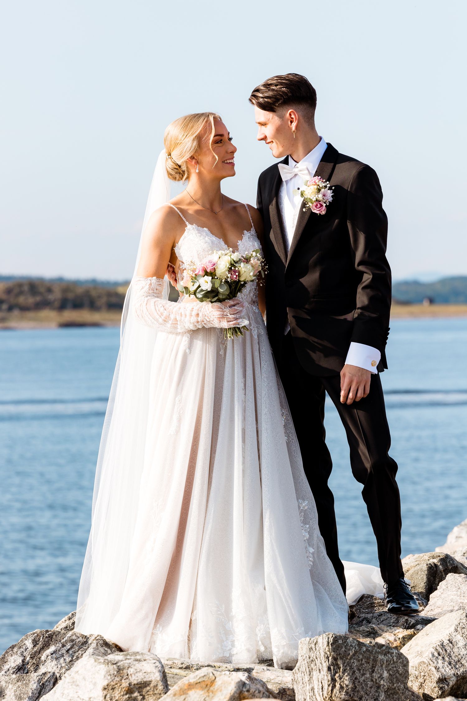 Newlyweds posing together on rocky coastline with water in background.