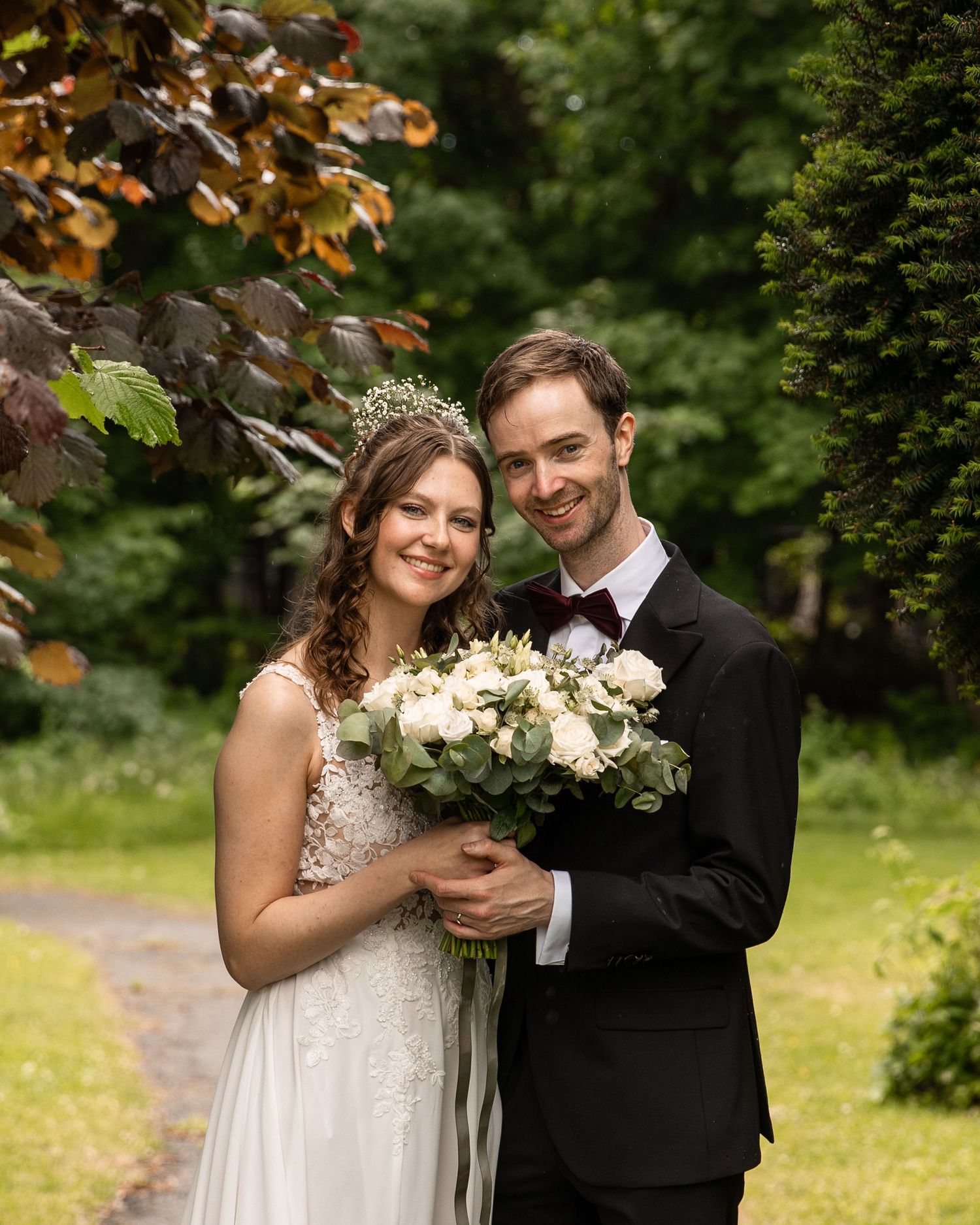 Bride in lace dress and groom in tuxedo share joyful moment with bouquet in garden.