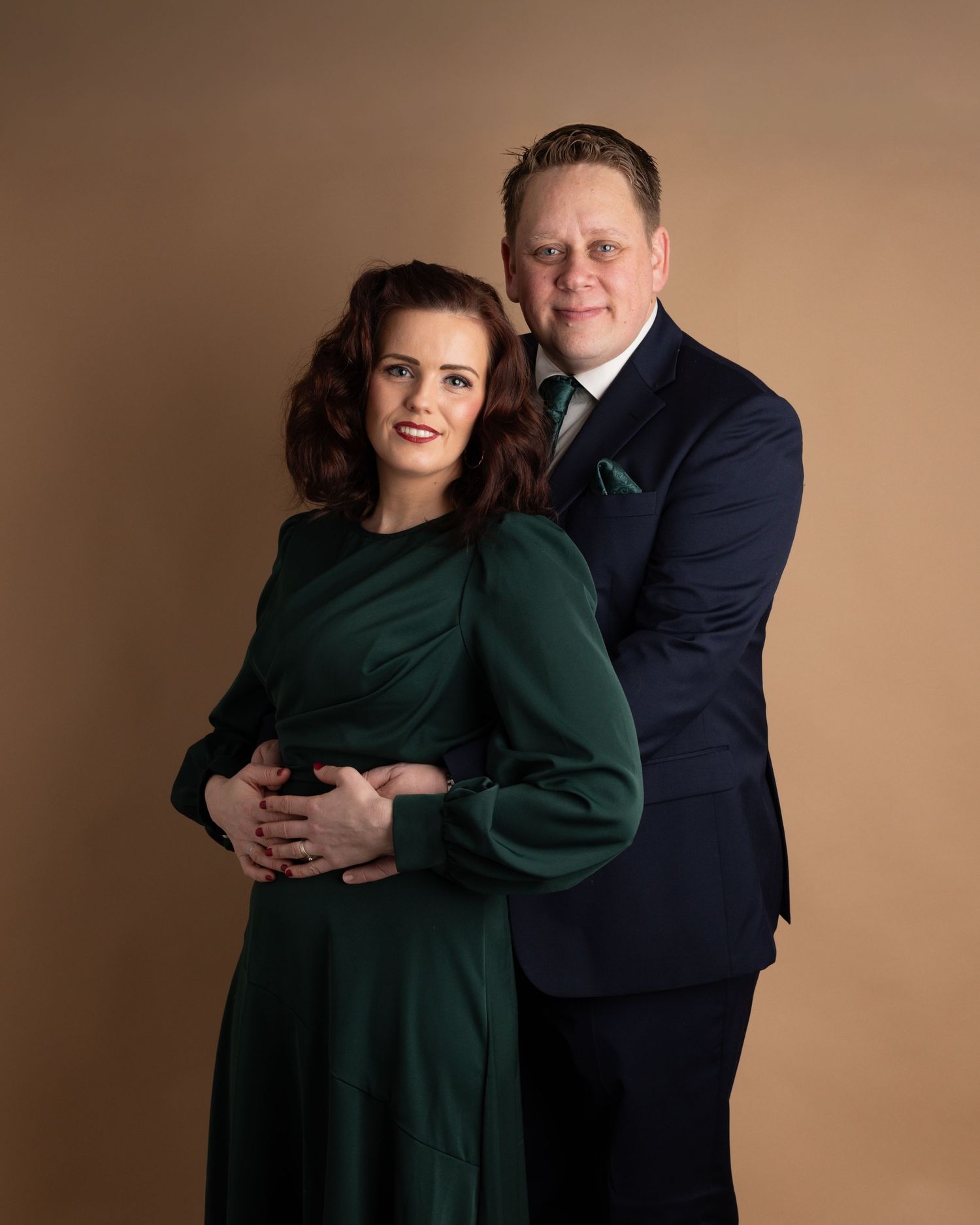 A couple poses together in a studio portrait, with a woman in emerald green dress and a man in navy suit against brown backdrop.