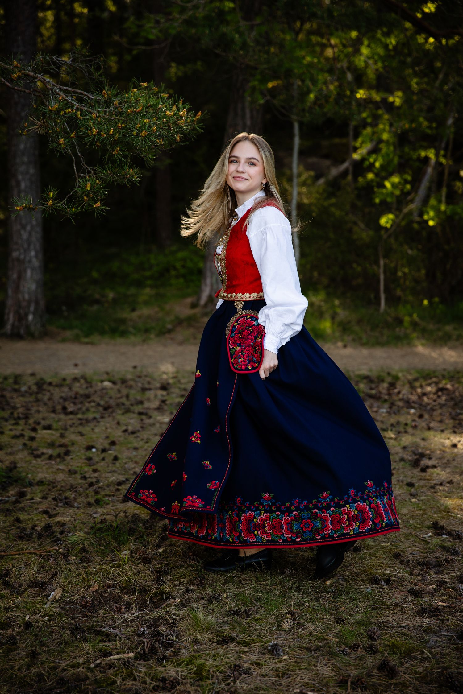 Traditional Norwegian folk dress with navy skirt and red embroidery twirling in forest setting.