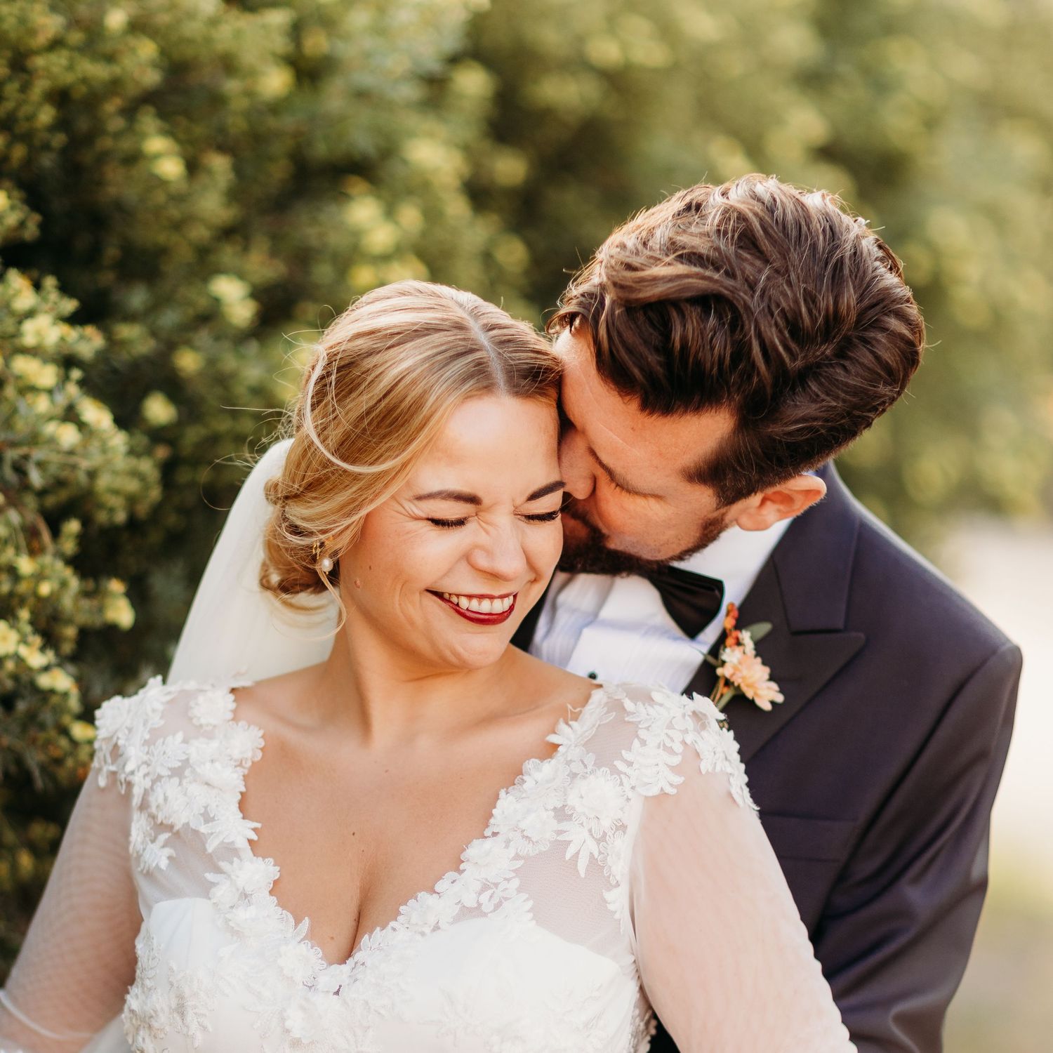 Intimate wedding moment captured with couple laughing against green foliage.