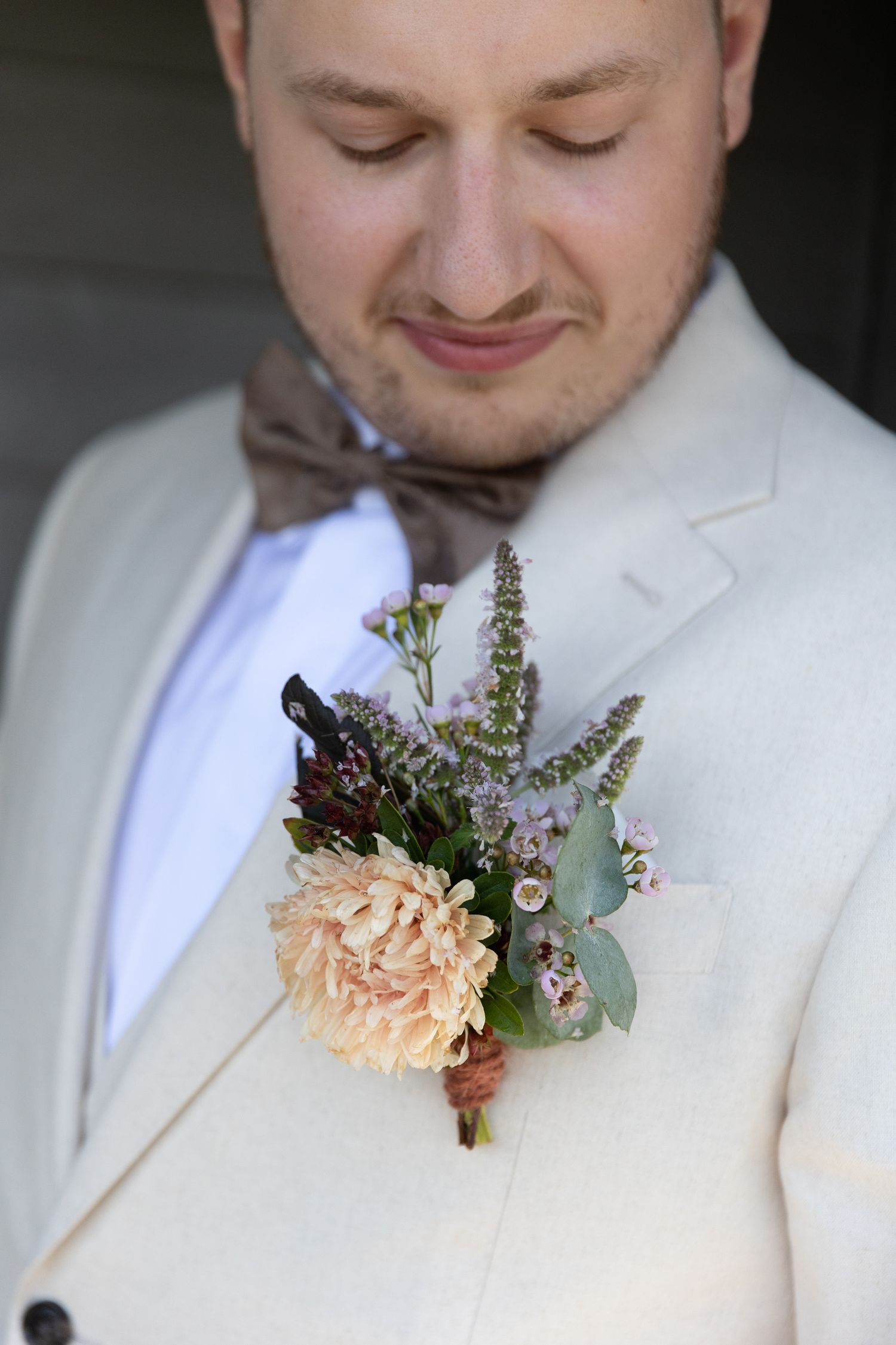 Close up of a boutonniere with peach carnation and purple flowers pinned to a cream suit jacket.