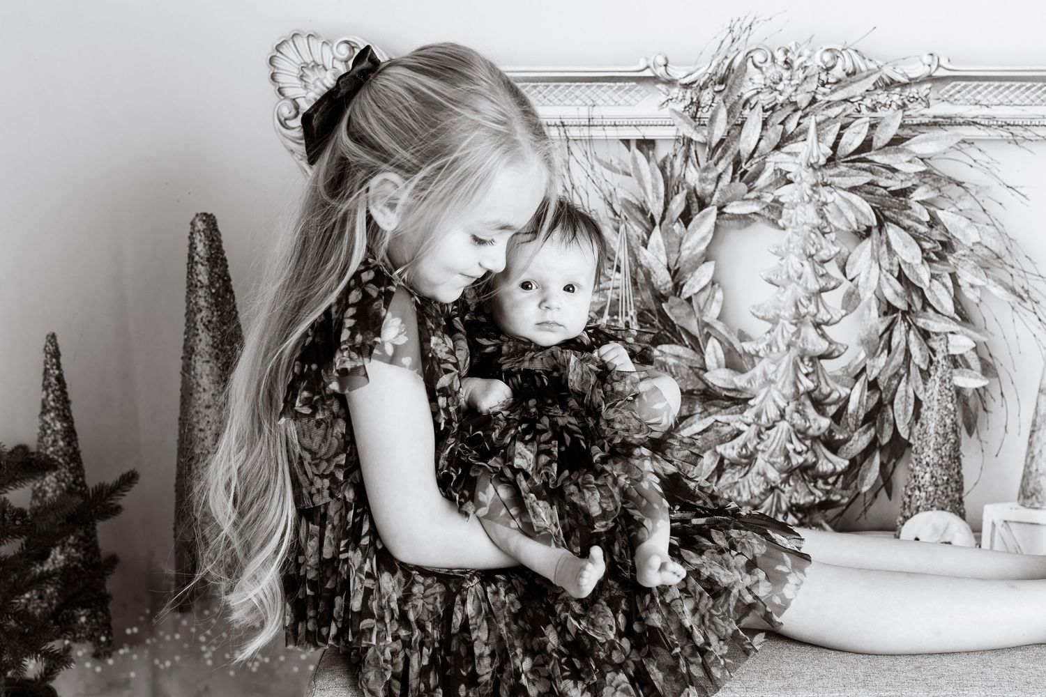 Black and white portrait of two children in matching floral dresses with holiday wreath backdrop.