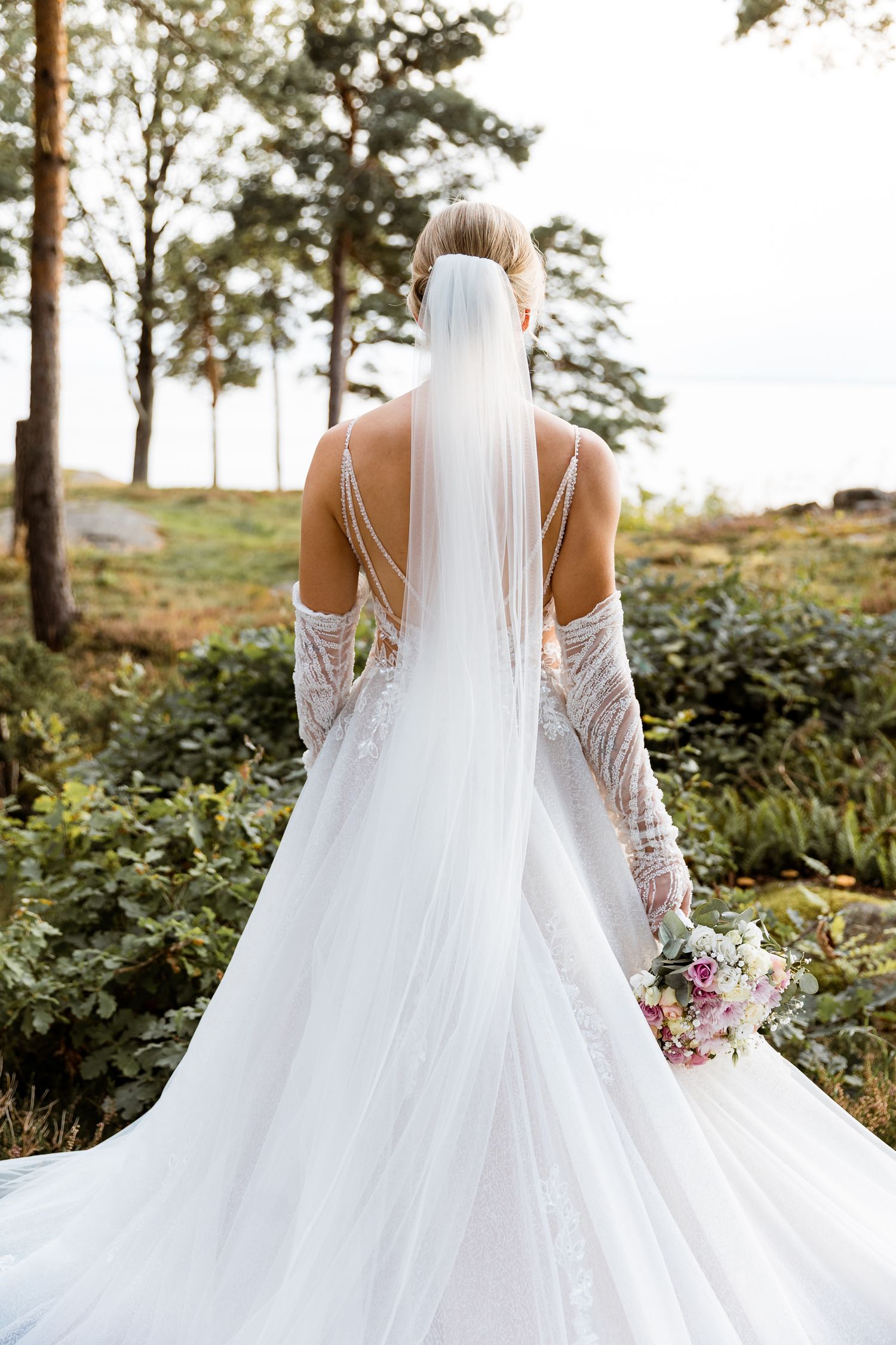 Elegant white wedding dress with flowing train and veil photographed from behind in a natural outdoor setting.