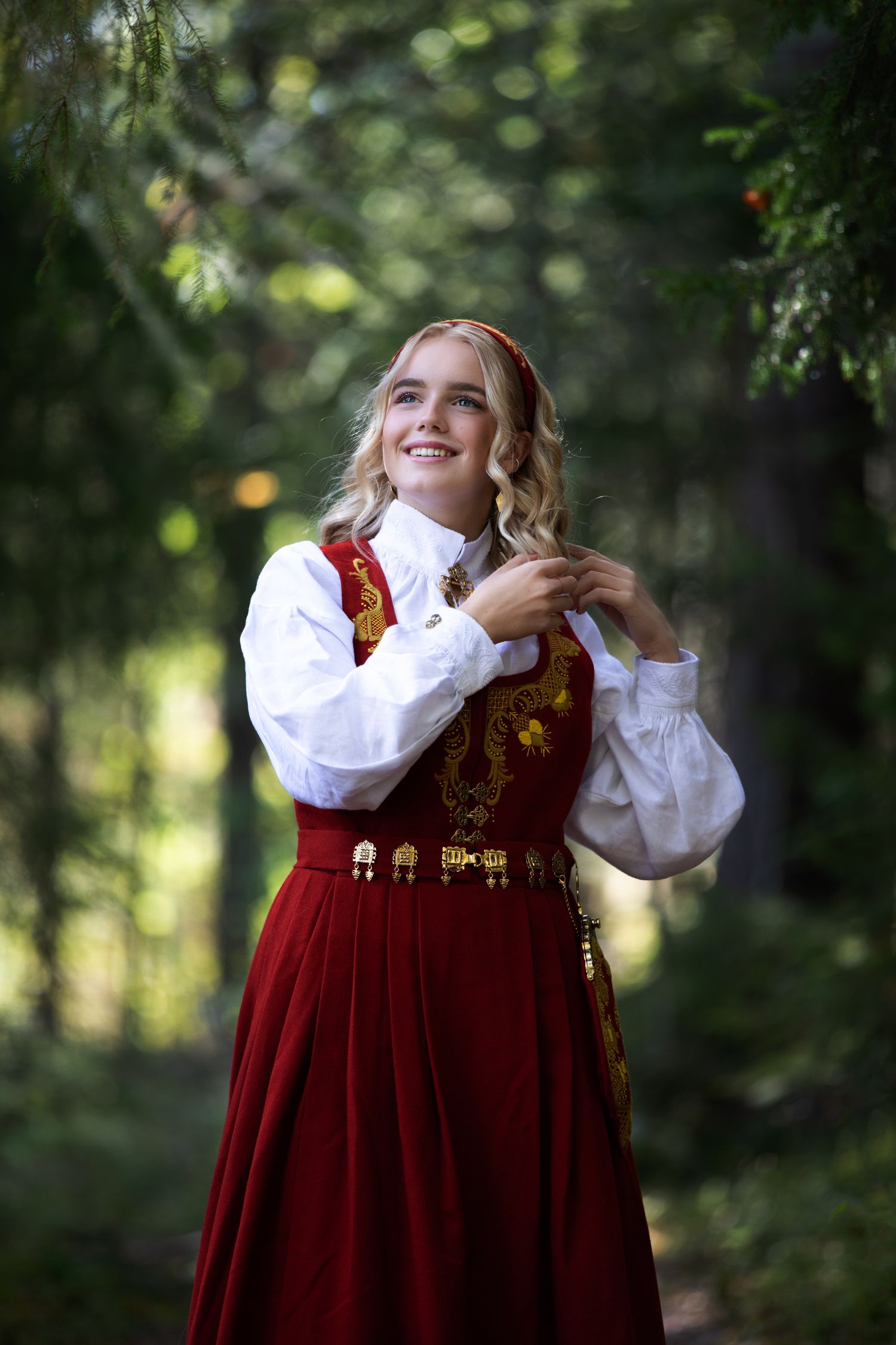 Red Norwegian bunad dress with gold embroidery shown in forest setting.