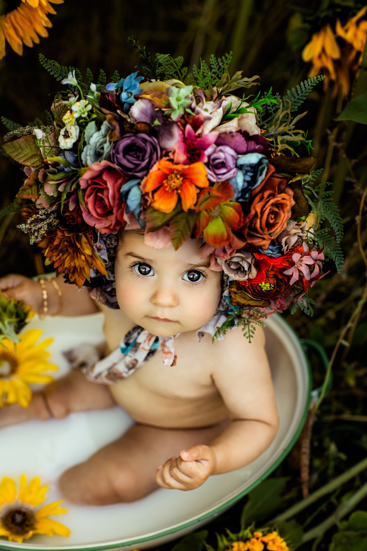 Infant wearing colorful floral crown lying on white surface with sunflowers in background.