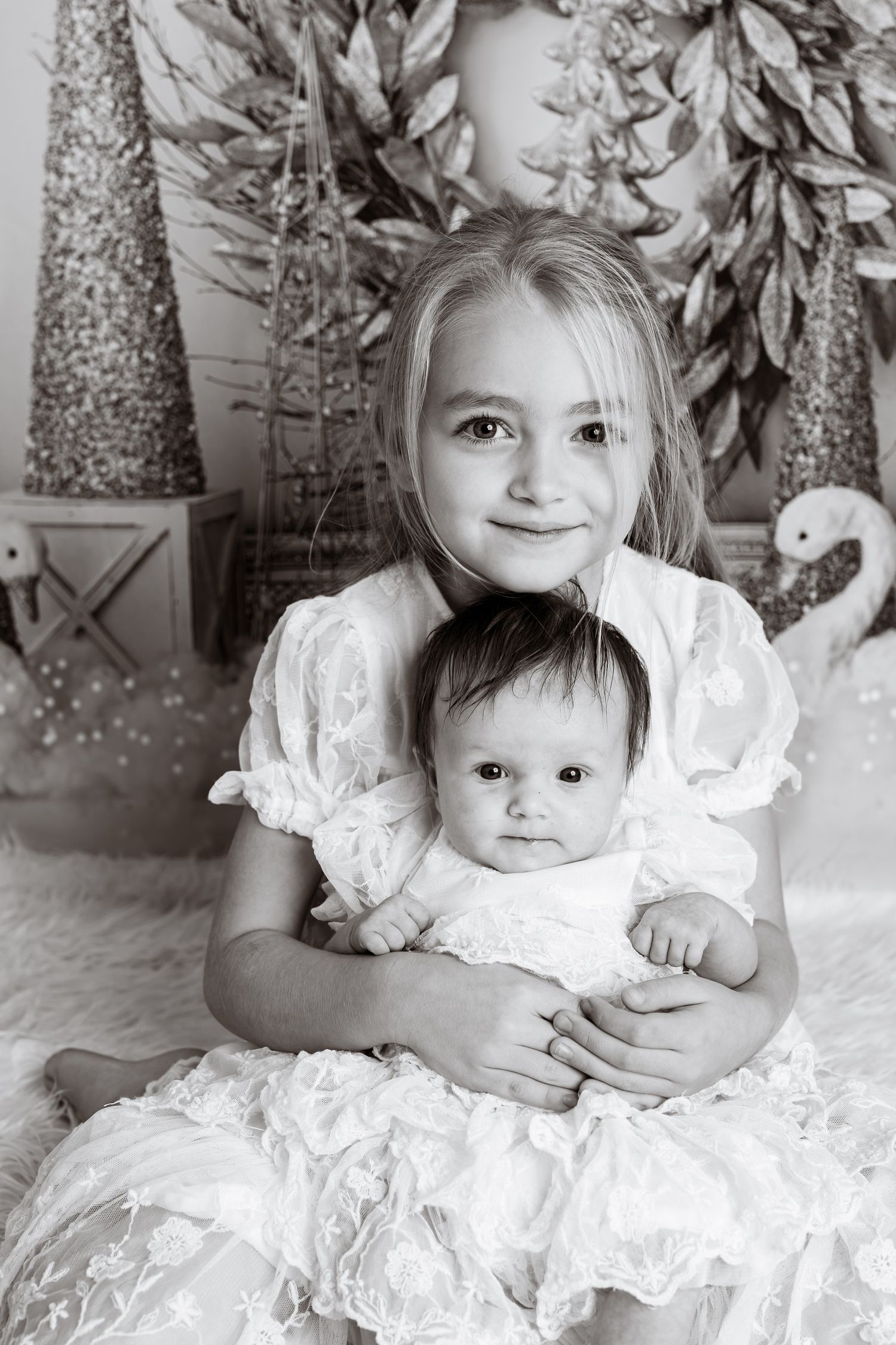 Black and white portrait of two siblings in white lacy dresses against a holiday backdrop with silver trees.