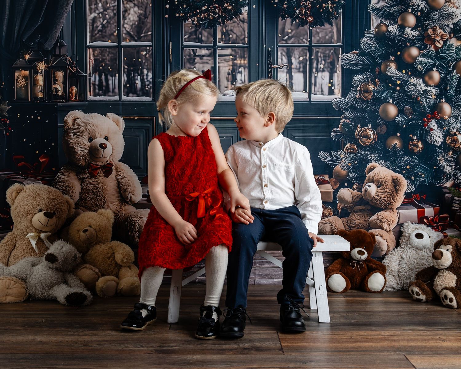 Two young children in holiday attire sit together surrounded by teddy bears and a decorated Christmas tree.