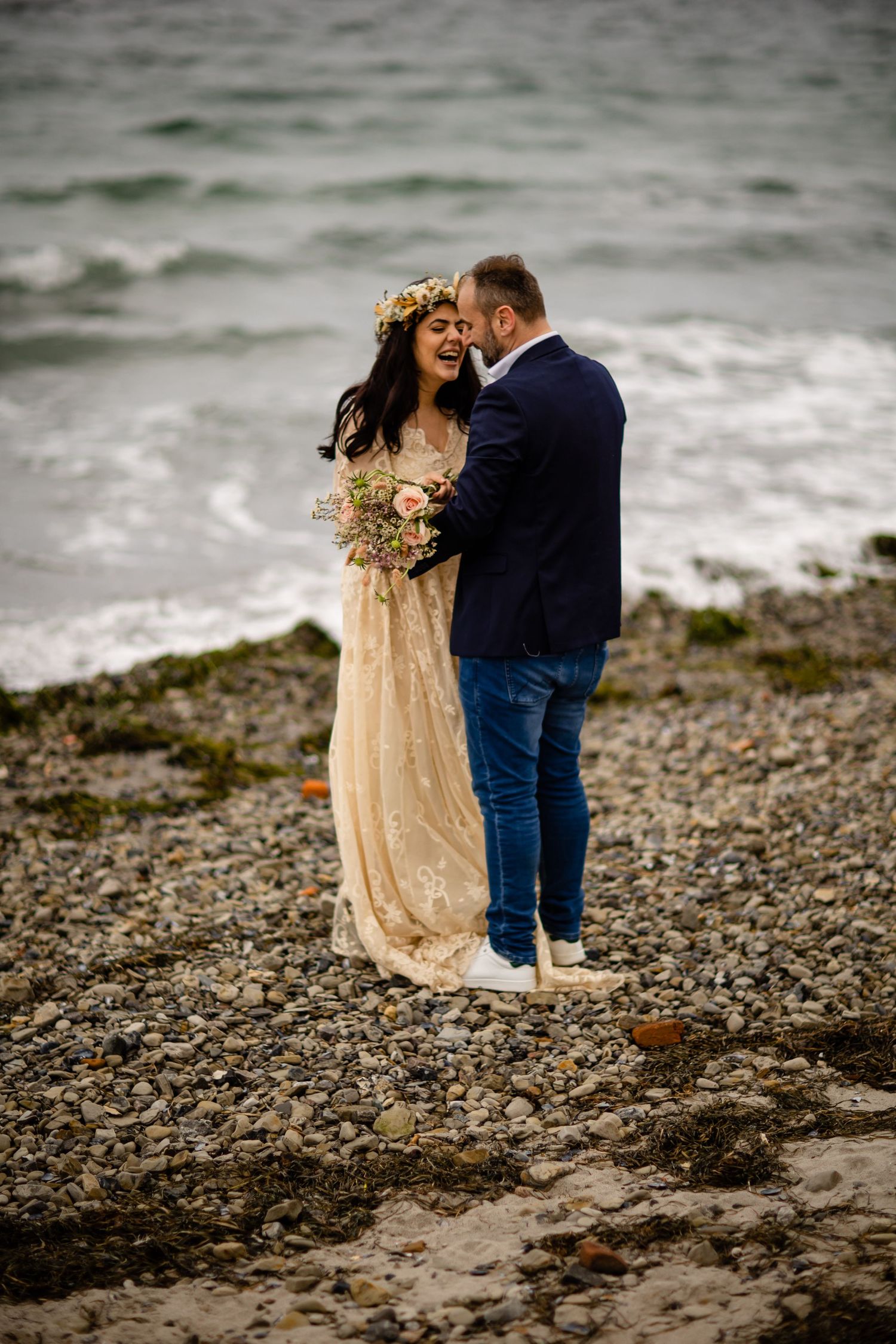 A romantic couple embraces on a rocky beach shore wearing a champagne dress and navy suit.