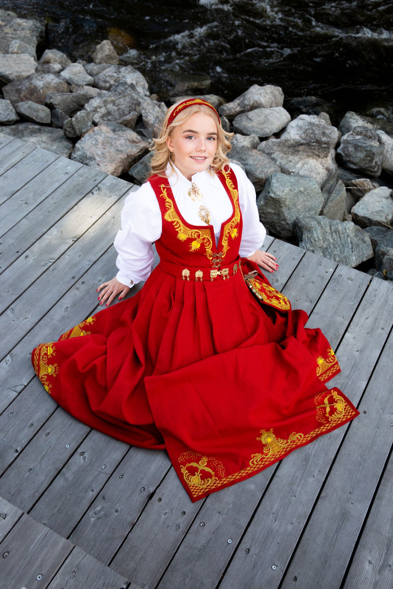 Traditional Norwegian bunad dress with red embroidered details displayed on wooden deck.
