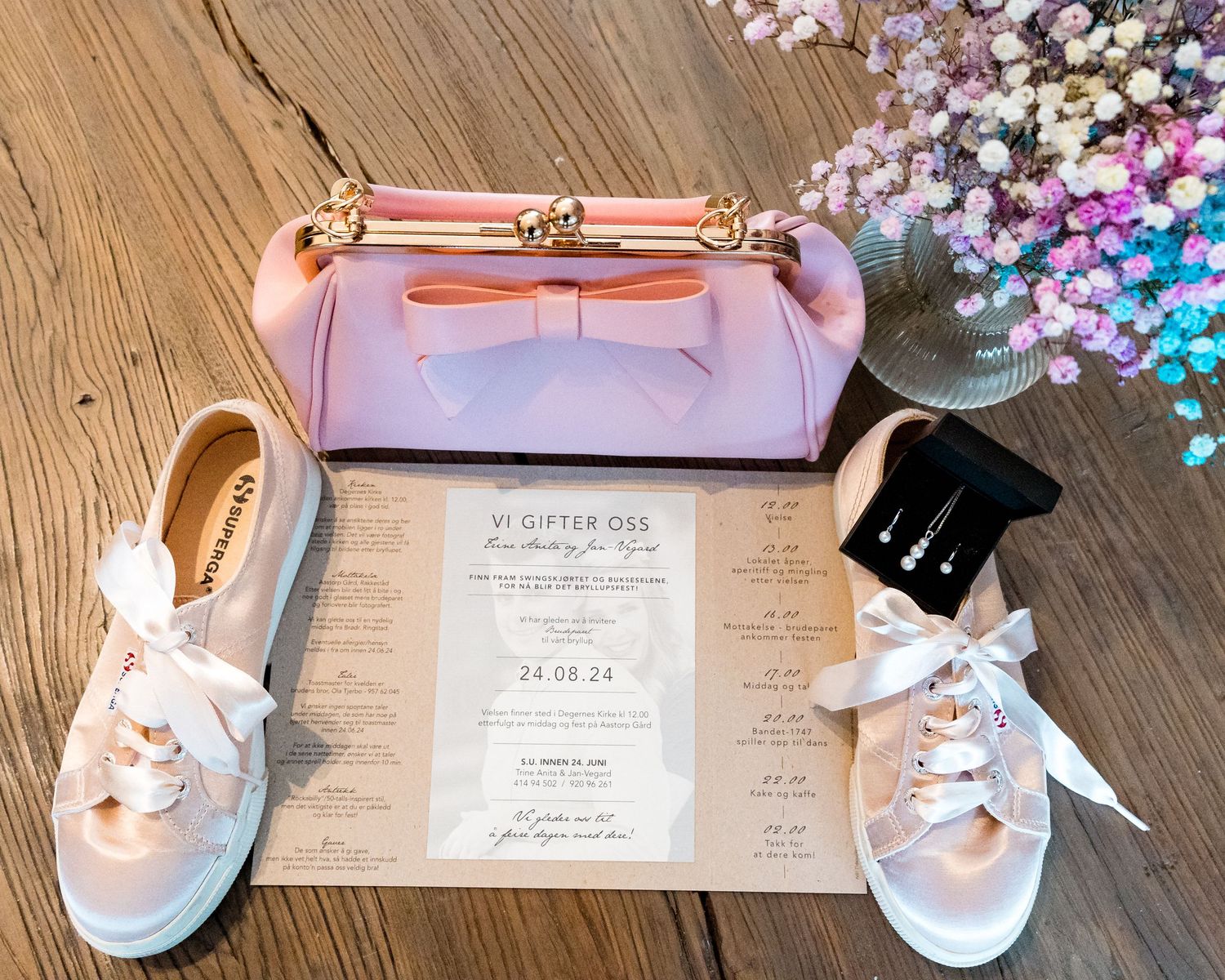 Wedding details flatlay with pink purse, bridal shoes and invitation on wooden surface.