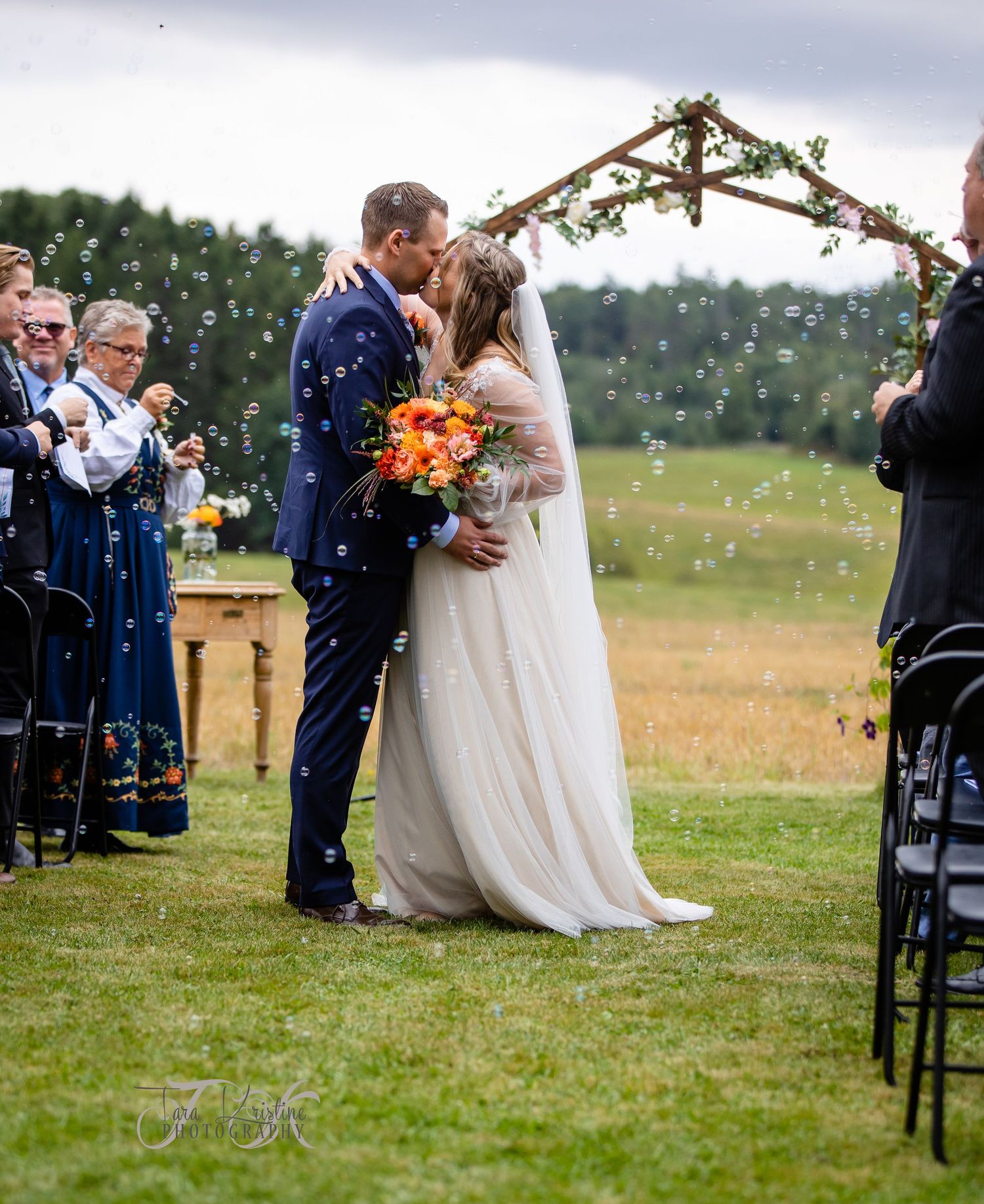 Outdoor wedding ceremony scene with guests seated on chairs in a grassy field.