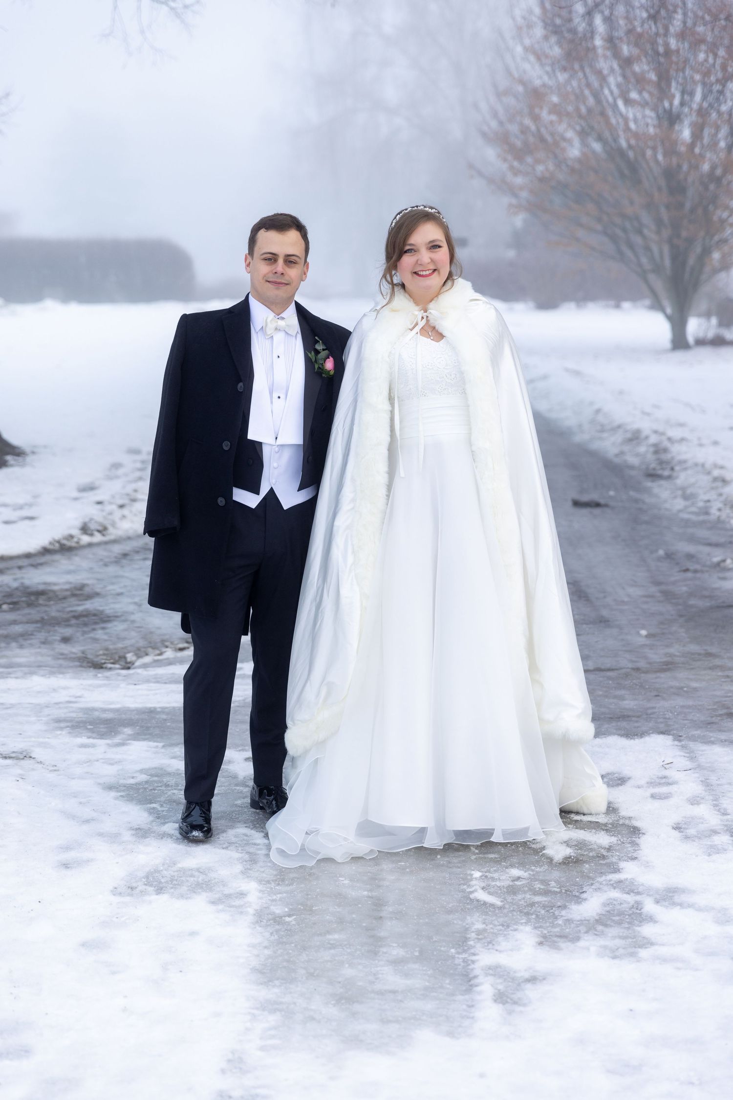 Vinterbryllup i Østfold: Brudepar i snødekt landskap, bruden bærer en hvit fuskepelskåpe foran et tåkelagt vinterlandskap ved Rygge kirke.