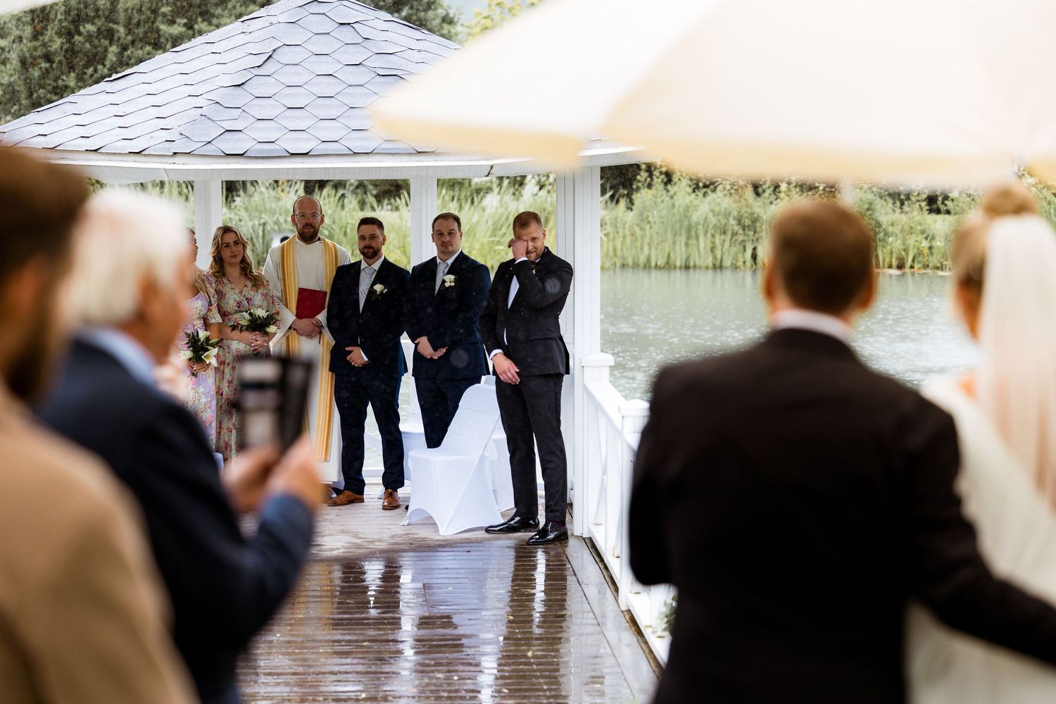 Wedding ceremony taking place under a white tent canopy on a wooden deck by a lake.