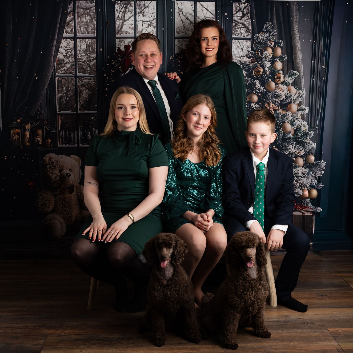 Family of five in matching green outfits pose for a festive Christmas portrait with a decorated tree and brown poodle.