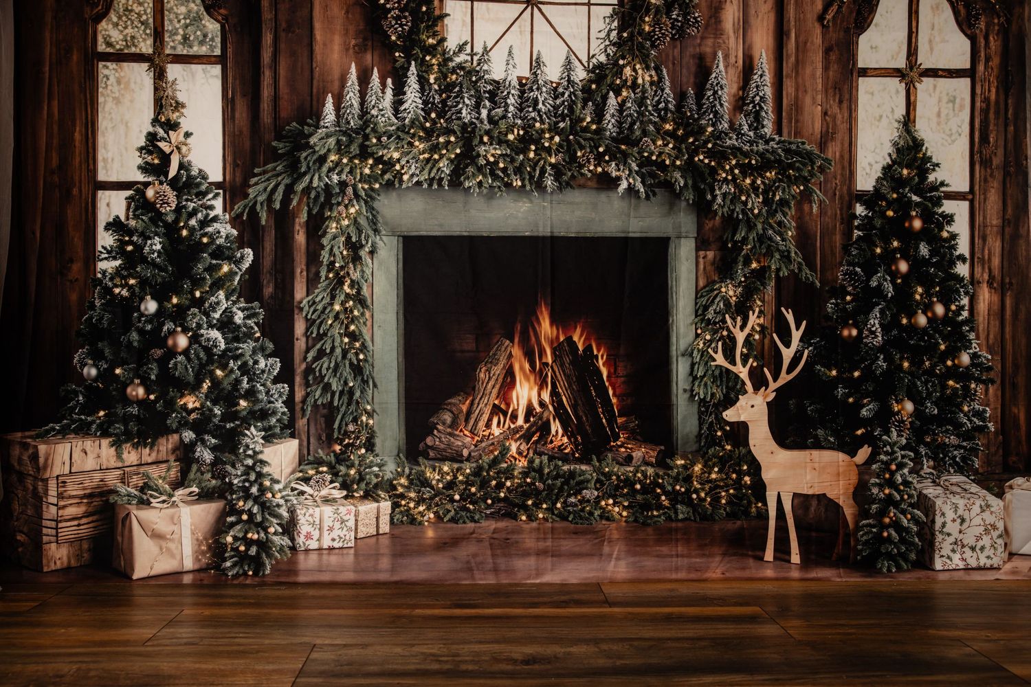 Cozy Christmas fireplace with a roaring fire, garland, decorated trees, wrapped gifts and a wooden reindeer figure on a rustic wood floor.