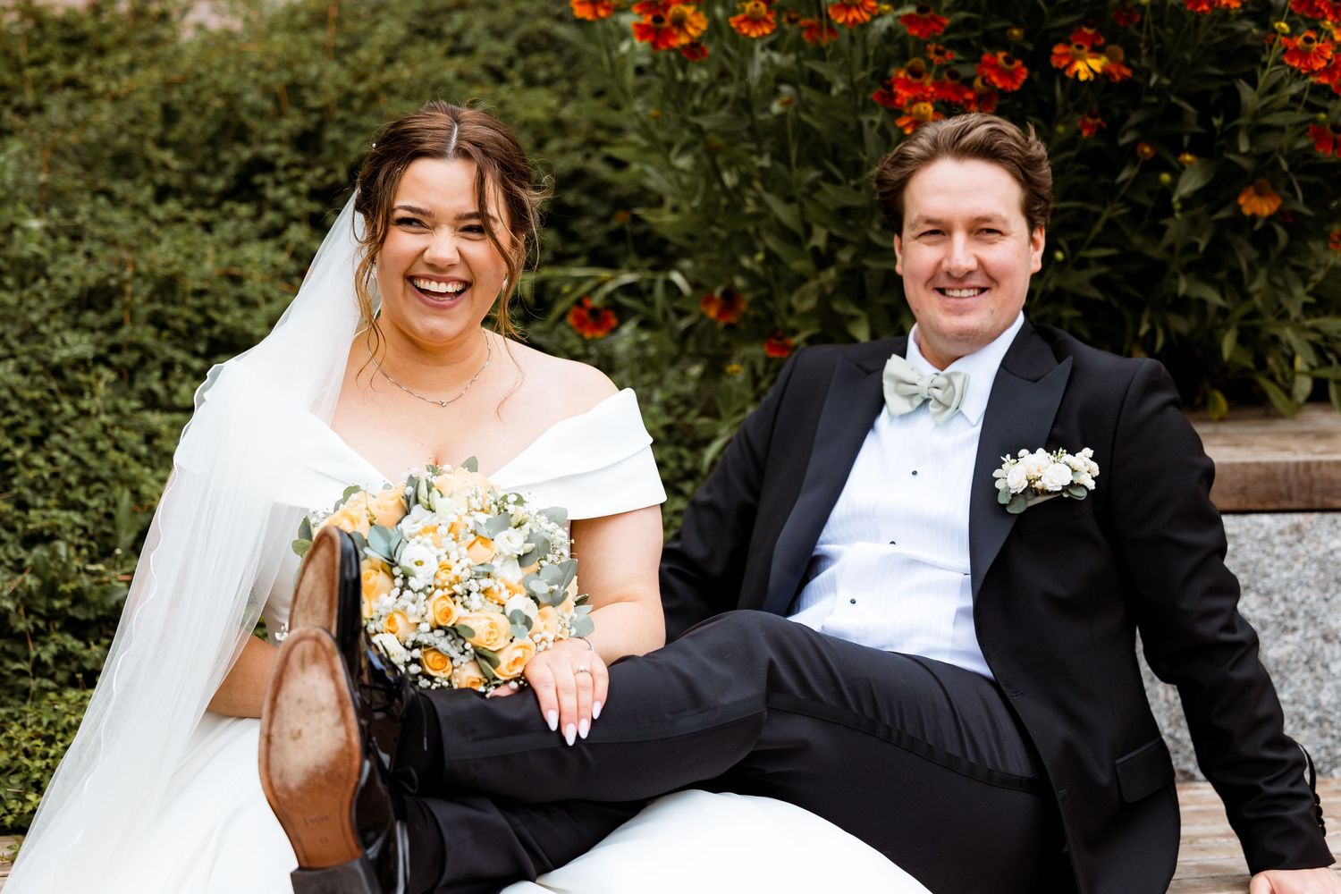 Wedding couple shares laughs on garden bench surrounded by red flowers.