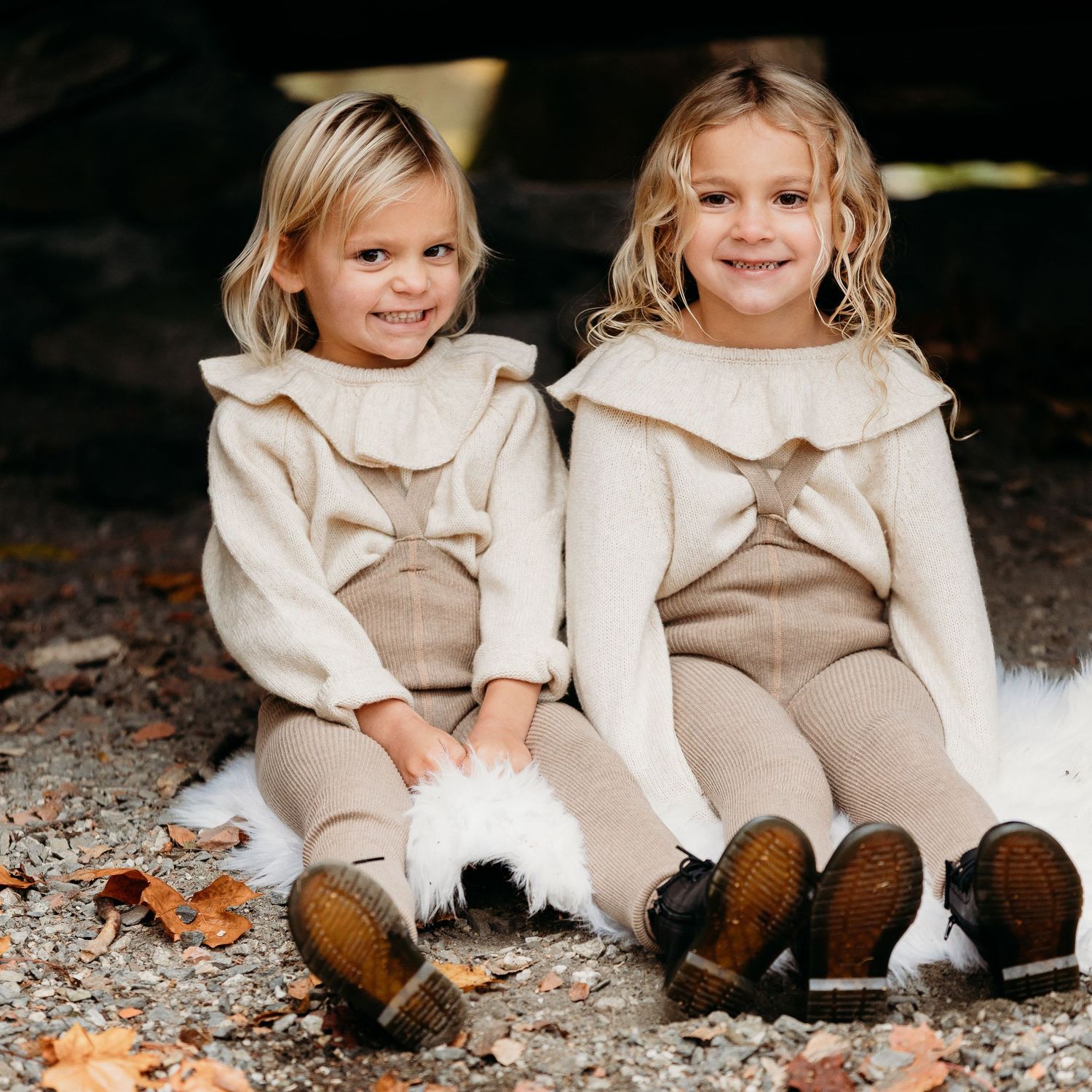 Two children in matching cream-colored outfits sitting together on autumn ground wearing boots.