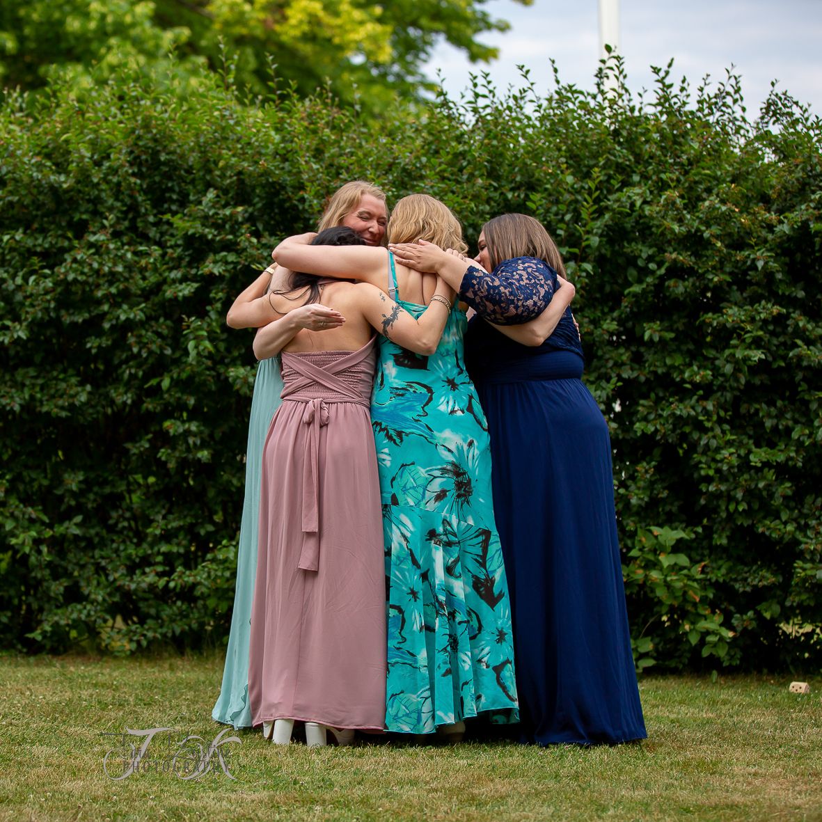 Three bridesmaids in colorful dresses pose together outdoors against green hedges.