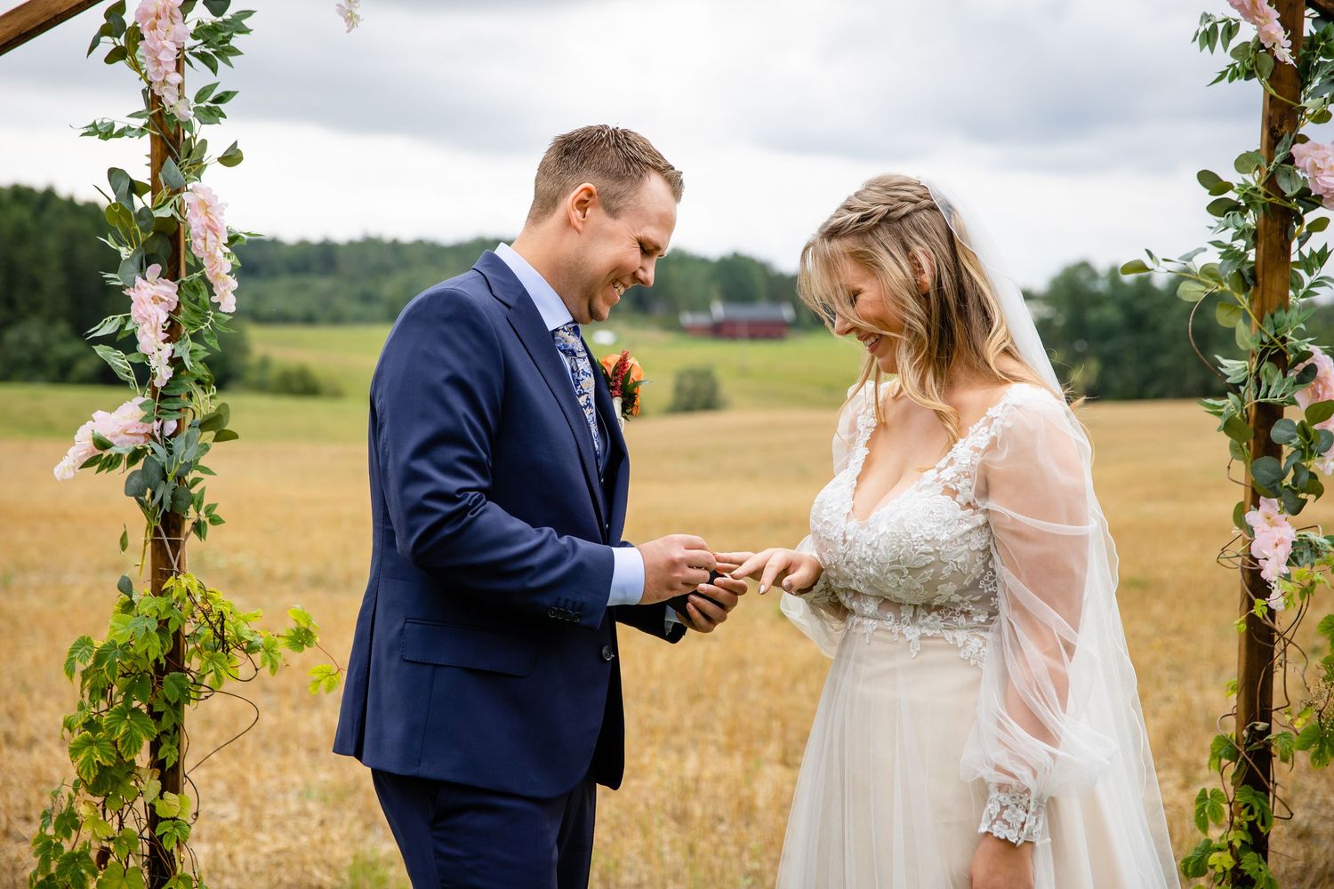 Outdoor wedding ceremony under floral arch in rustic field setting.