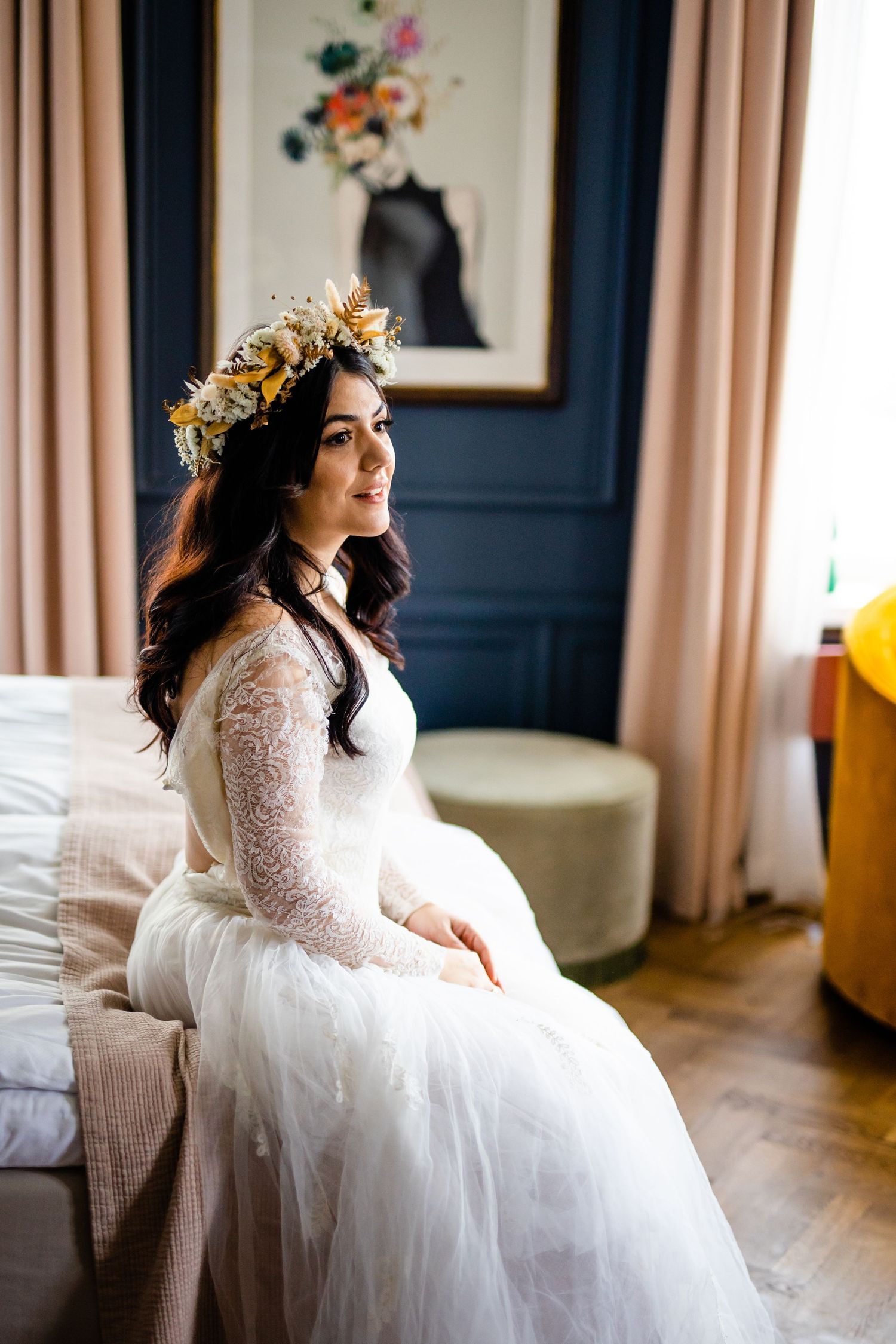 Bride in lace wedding dress sitting elegantly in a room with floral artwork.