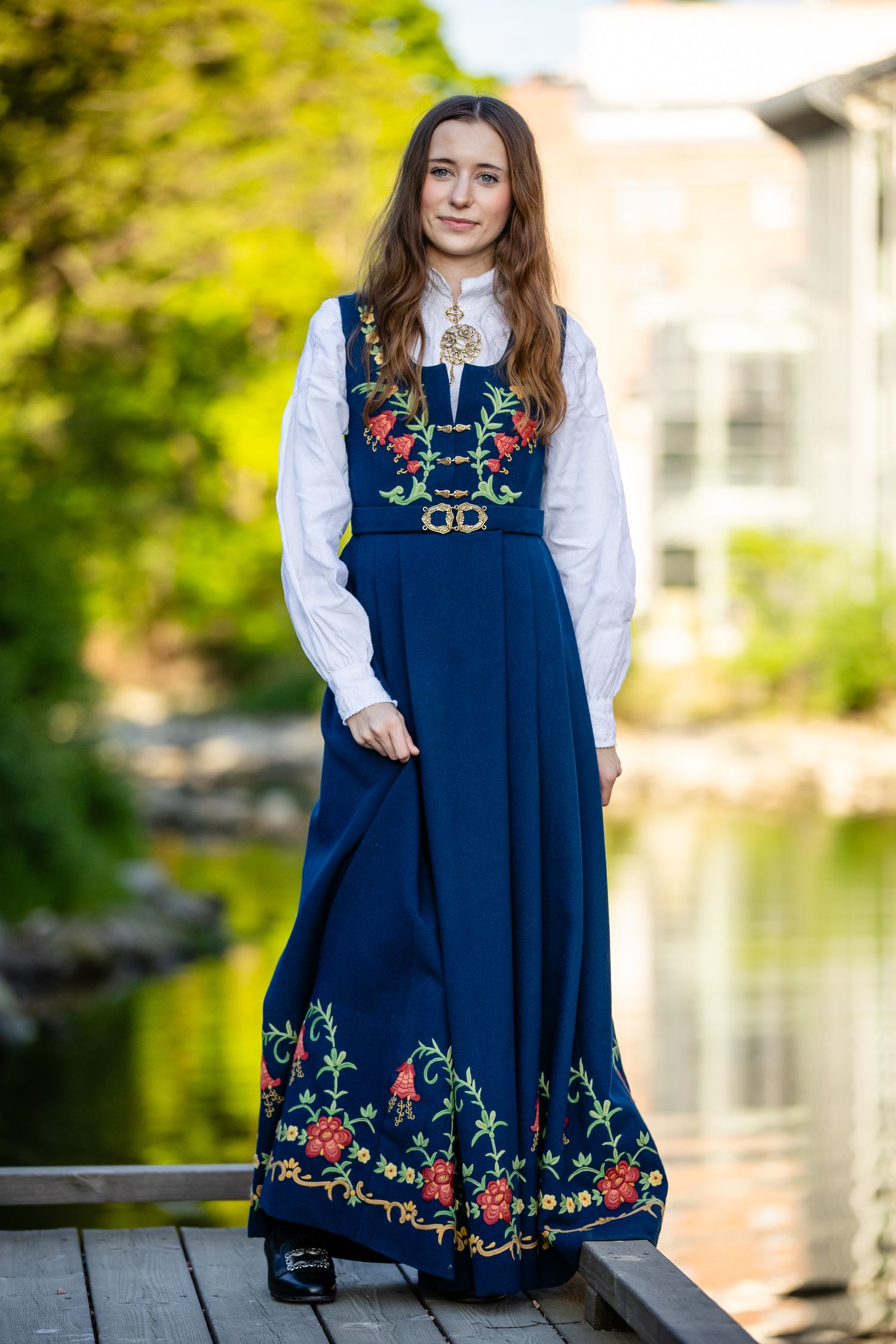 Person wearing navy folk dress with floral embroidery poses near water in natural setting.