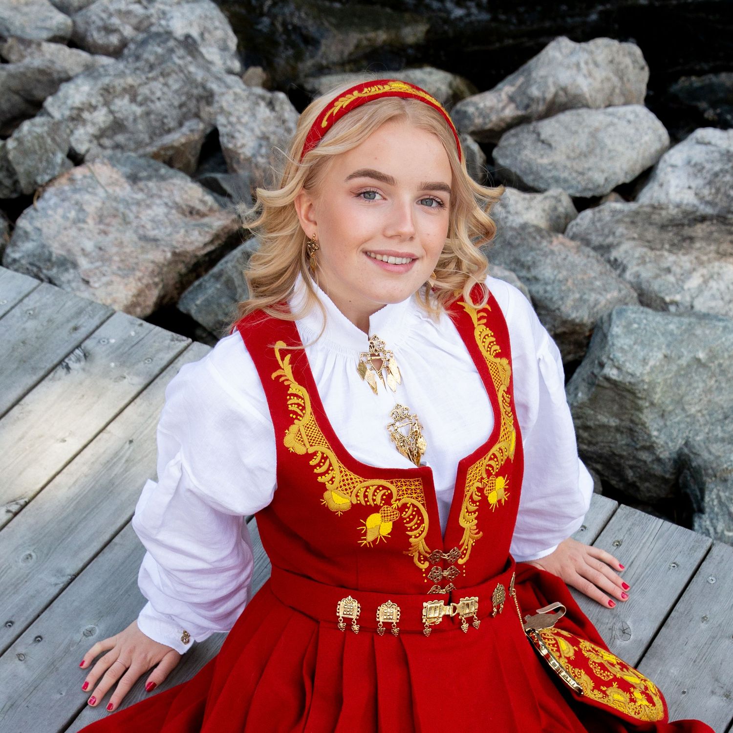 A portrait in traditional Norwegian bunad with red and gold embroidery sitting on wooden deck by rocks.