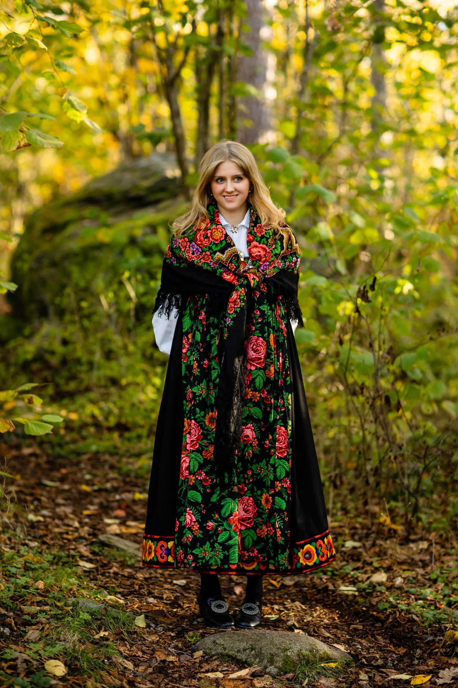 Traditional floral folk dress with vibrant red roses on black fabric photographed in autumn forest setting.