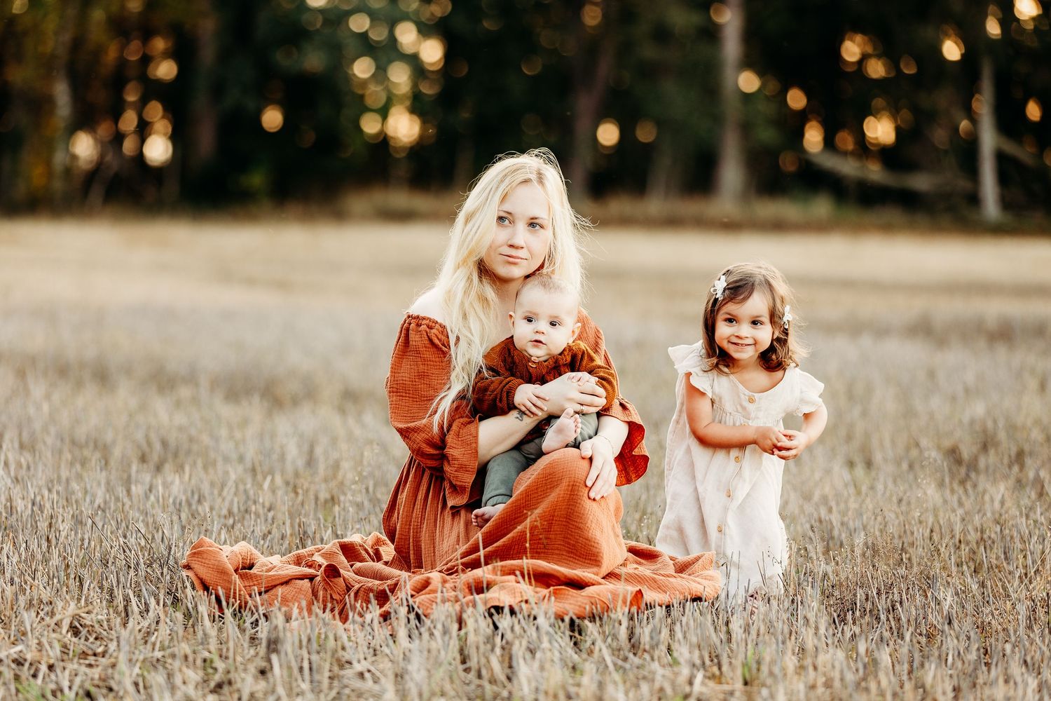Barnefotograf Moss: Mor sitter på en åker og holder sin lille baby, mens en smilende storesøster står ved siden av i det gylne kveldslys