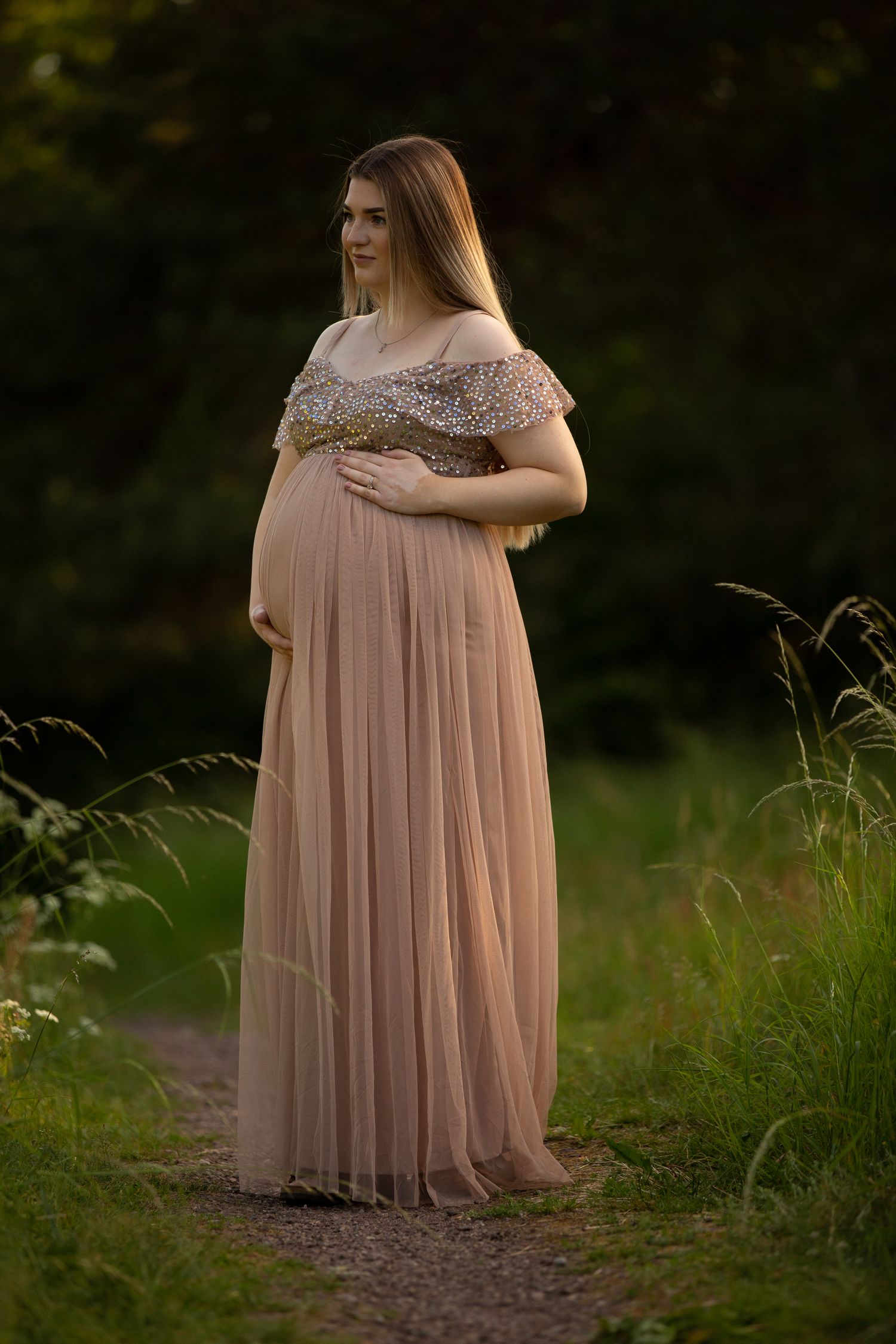 Maternity portrait in flowing pink dress surrounded by tall grass at sunset.