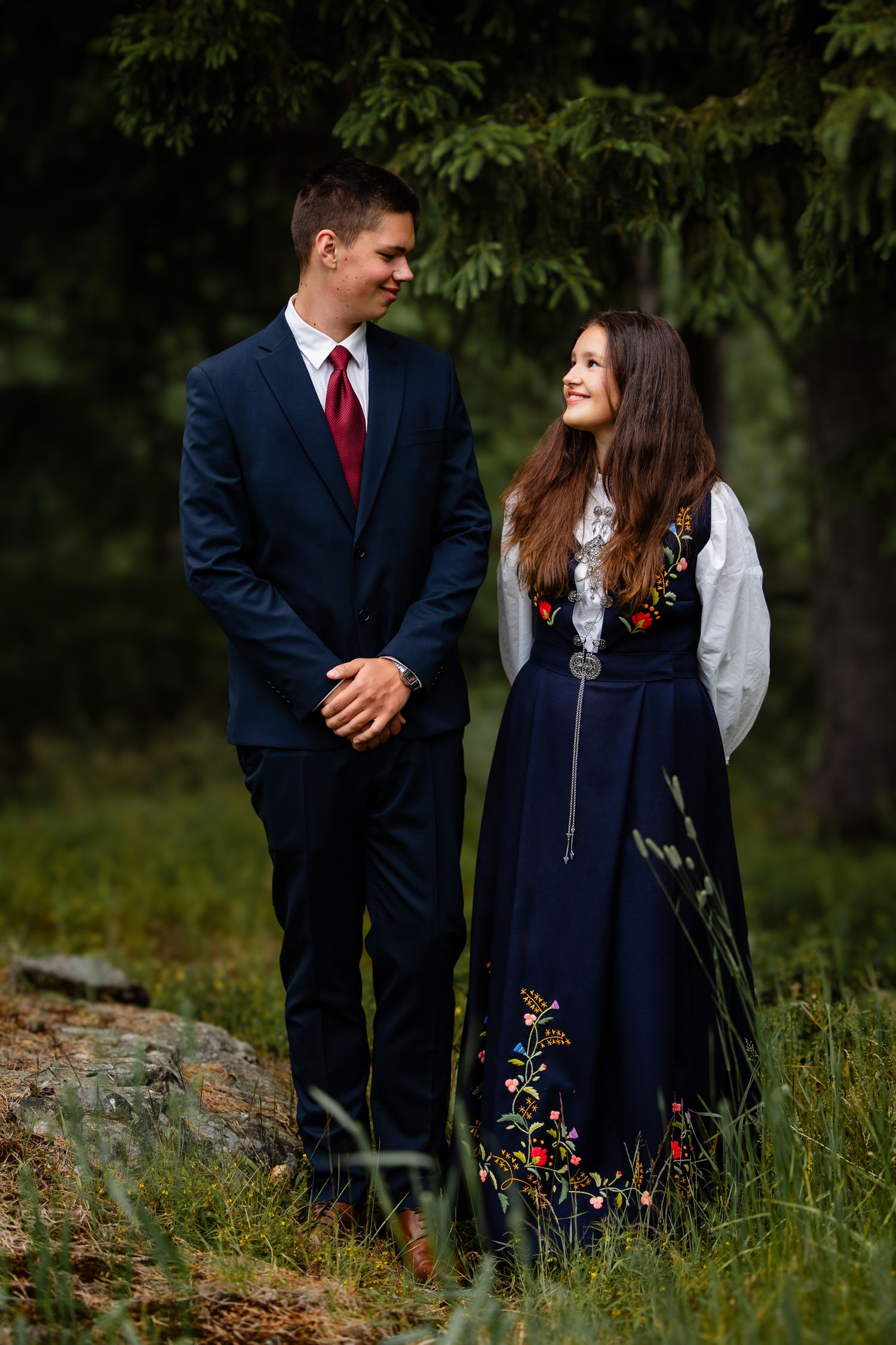 A couple stands outdoors wearing formal attire, with him in a navy suit and her in an embroidered navy dress.