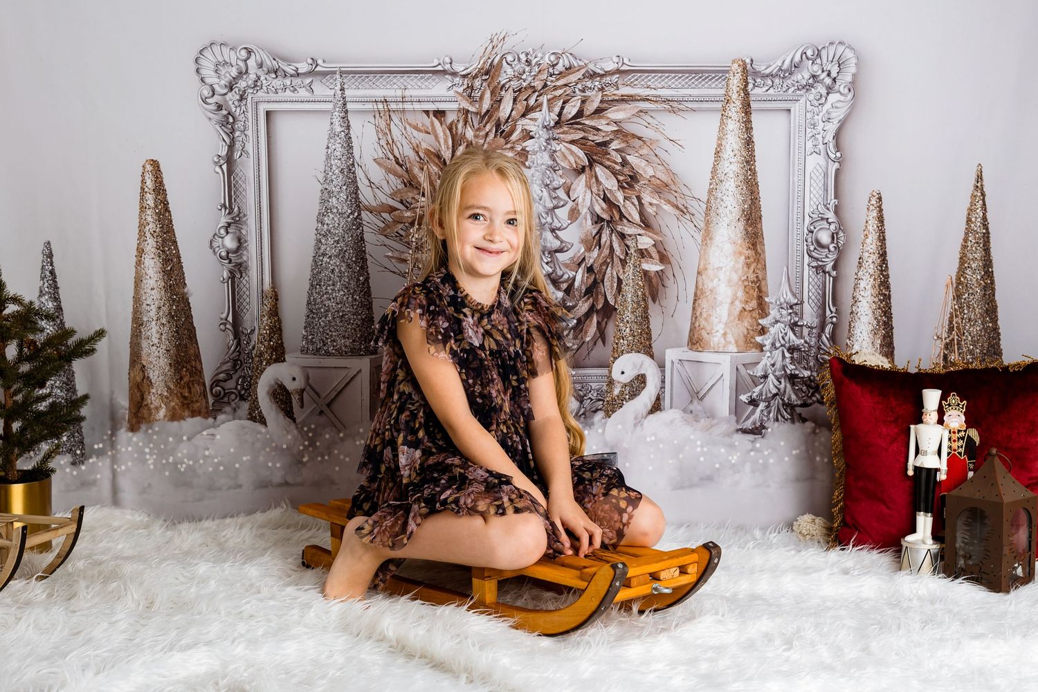 Child in dark dress poses on wooden bench with holiday decor and metallic trees.
