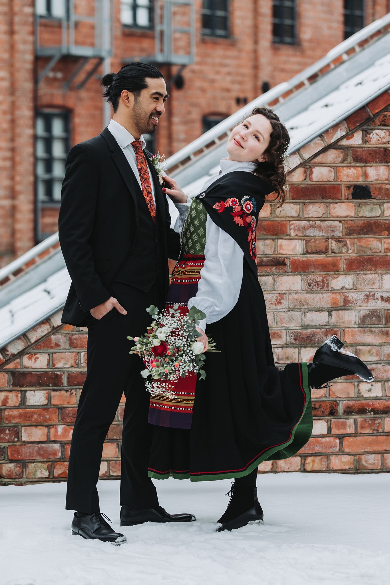 A couple dances together in formal wear against a snowy brick wall.