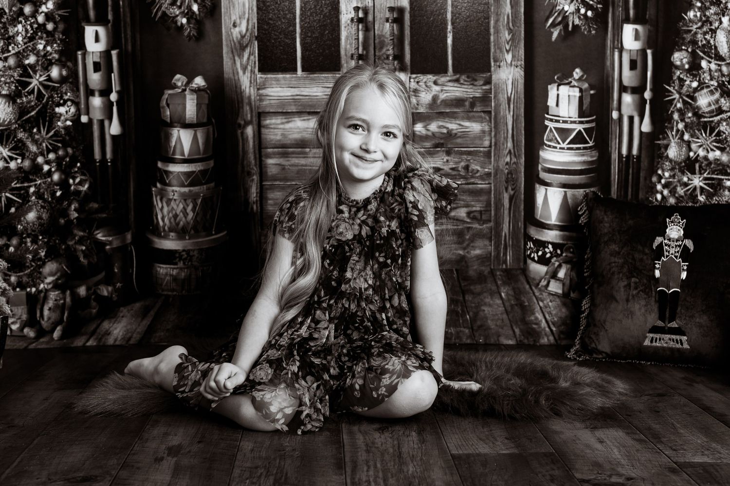 A black and white portrait of a child in a floral dress sitting on wooden floor with rustic barrels and decor.
