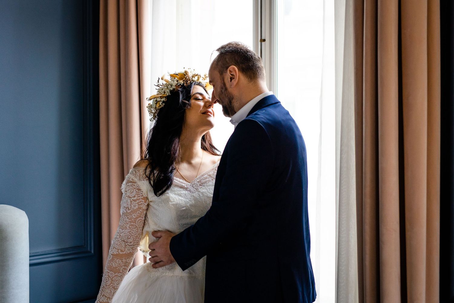 Wedding couple shares tender moment near window with soft natural light.